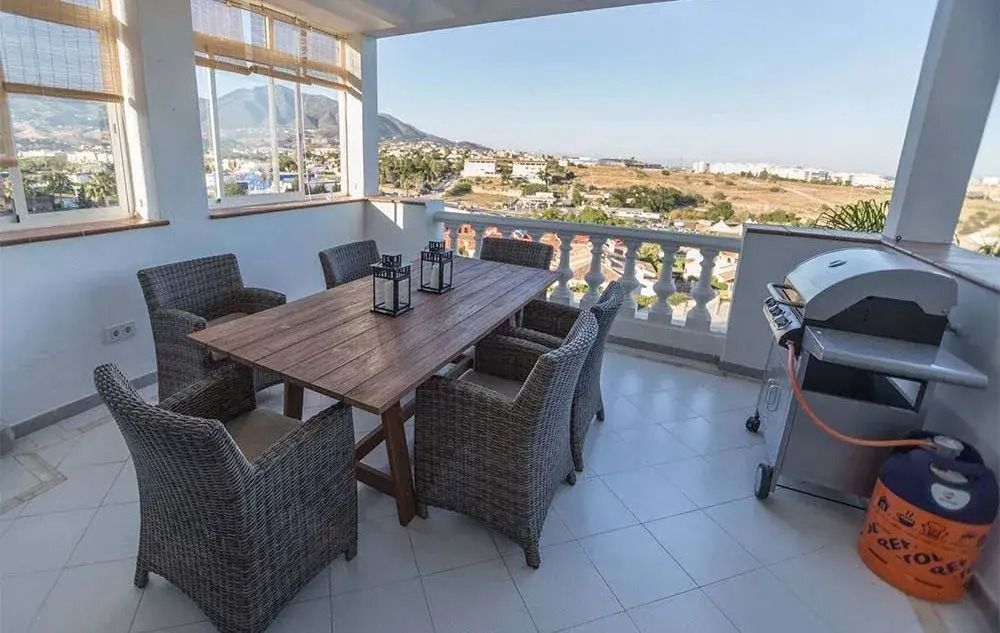 Patio dining area with wicker chairs, wooden table, and a grill overlooking a coastal view