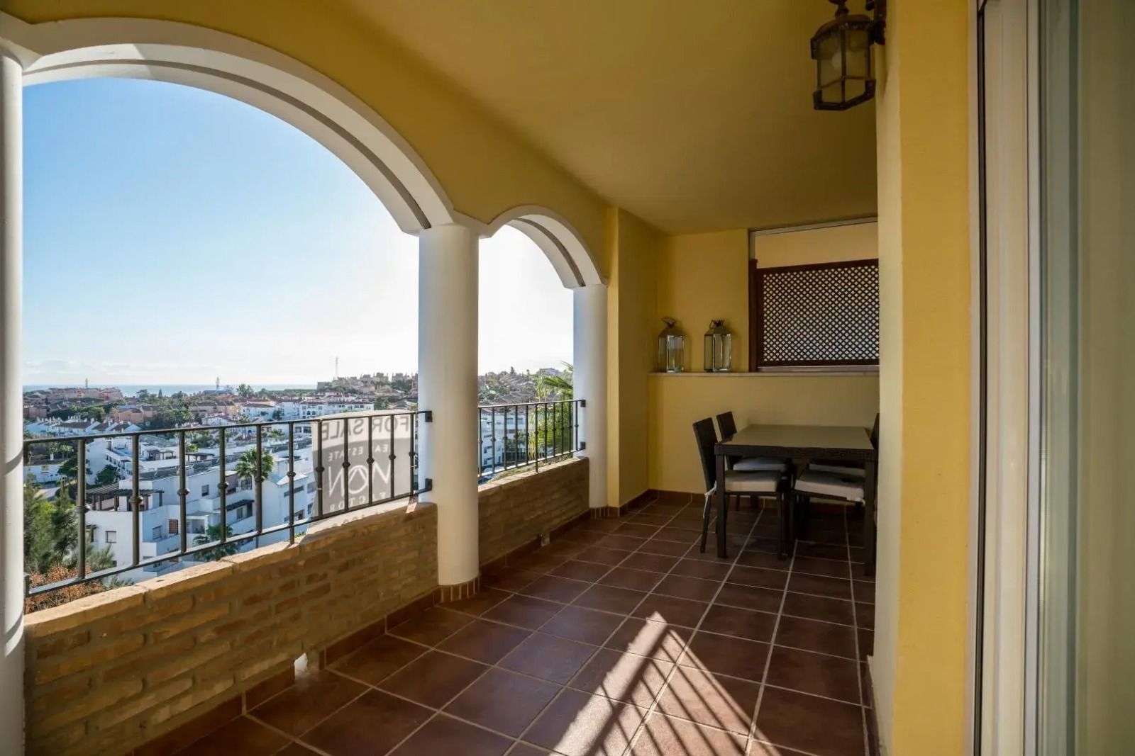 Covered balcony with tiled floor, arched openings, and a city and water view at sunset