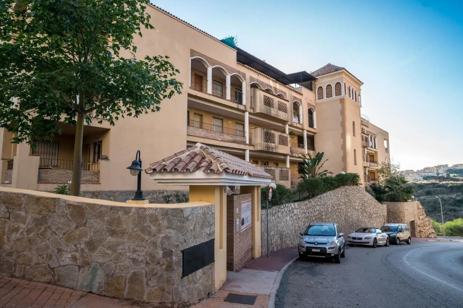 Stone apartment building with balconies on a curving hill road, with parked cars below.