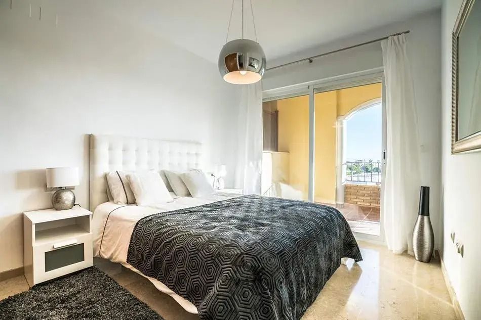 Bright bedroom with white furniture, dark bedspread, large window, and sliding glass door to balcony
