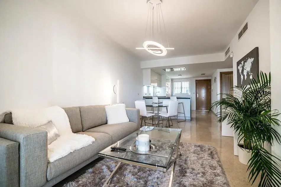 Bright modern living room with gray sofa, glass coffee table, rug, and open kitchen in the background