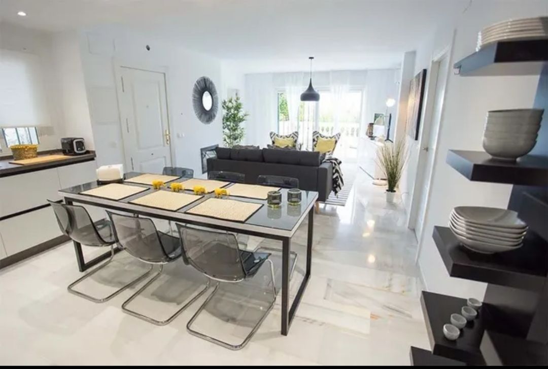 Modern open-plan dining and living room with a glass table, black chairs, and a gray sofa in bright daylight