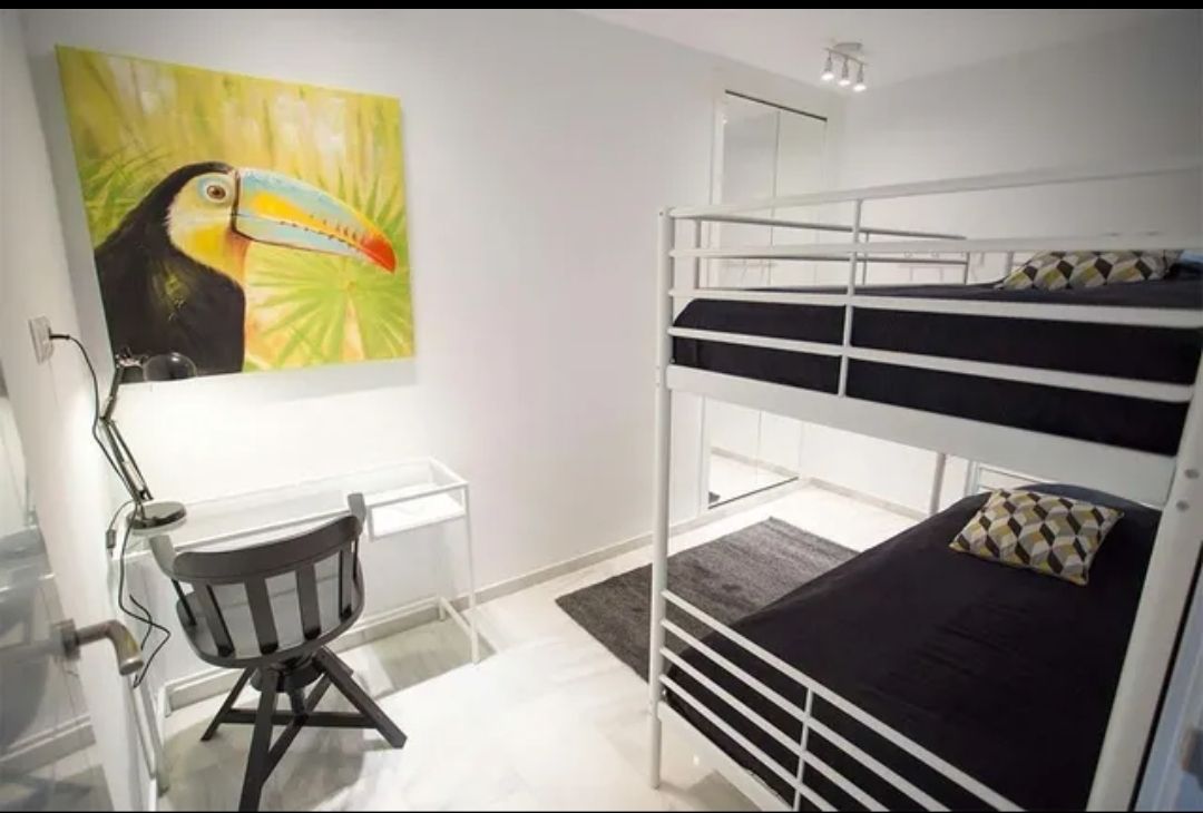 Small white bedroom with bunk bed, desk, chair, and toucan painting on the wall.
