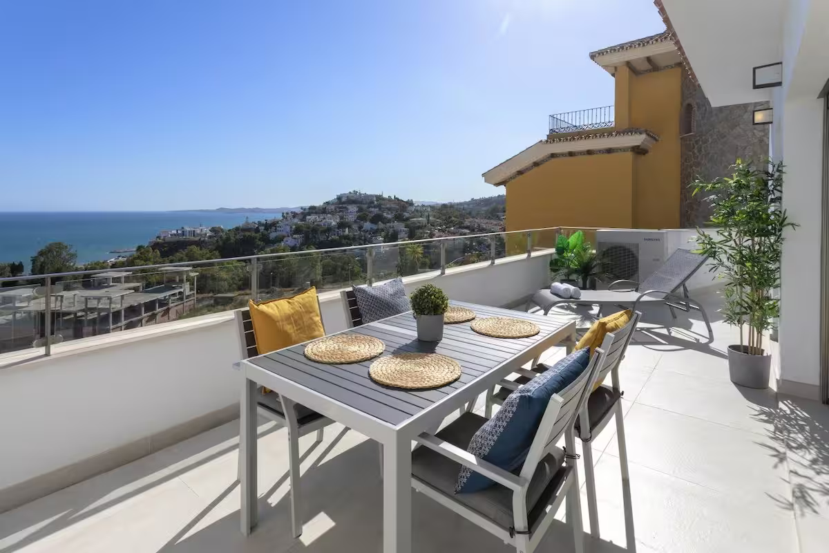 Sunny rooftop terrace with dining table, blue chairs, potted plants, and sea view