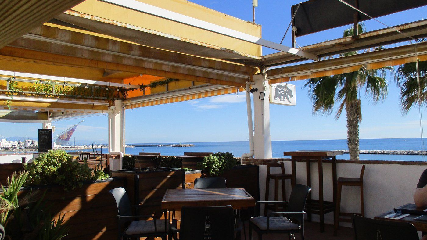 Shaded seaside café terrace with tables, chairs, and ocean view under yellow awnings