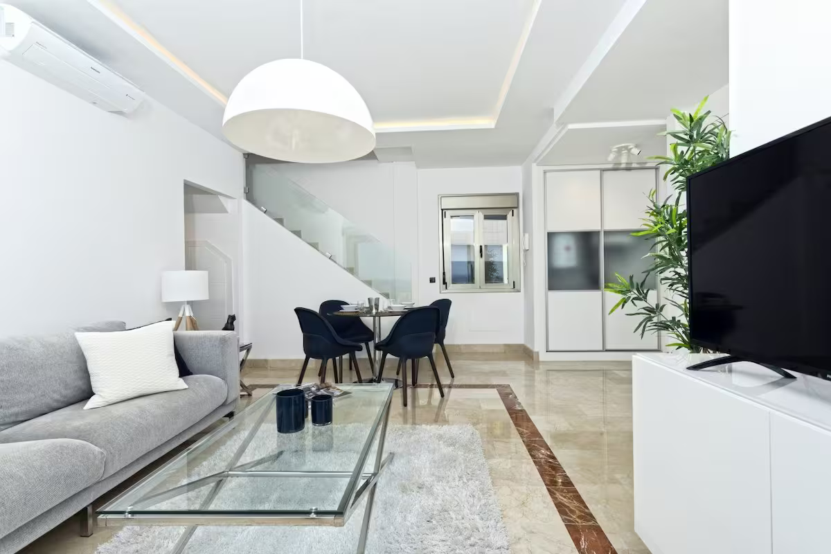Bright modern living room with gray sofa, glass coffee table, dining area, and large TV.