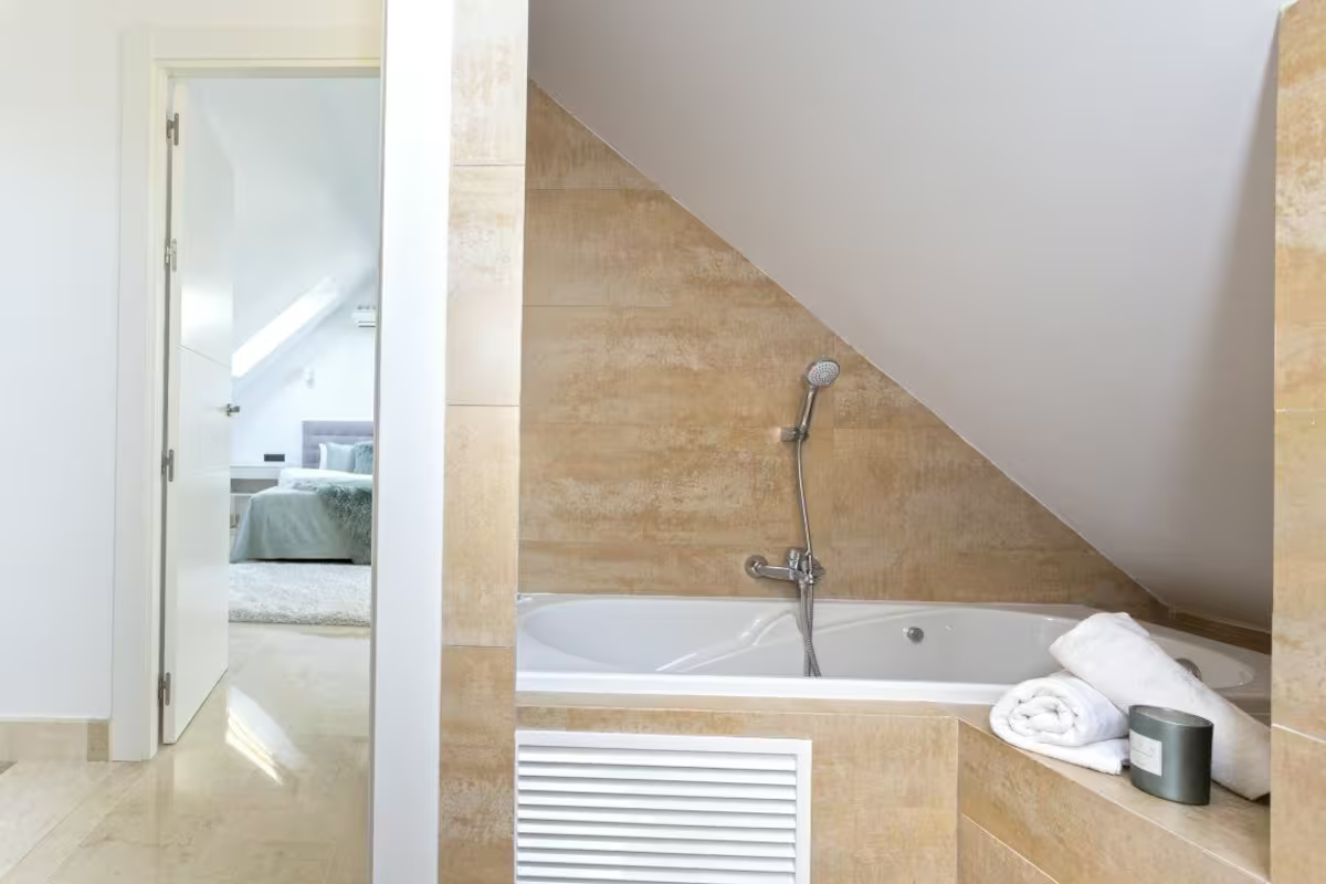 Modern bathroom with built-in bathtub, beige tile, sloped ceiling, and towels on the tub edge
