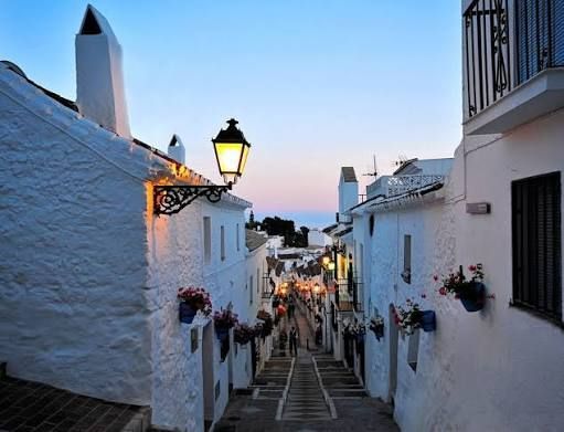 Narrow whitewashed street with lanterns and flower pots leading to a sunset-lit town center