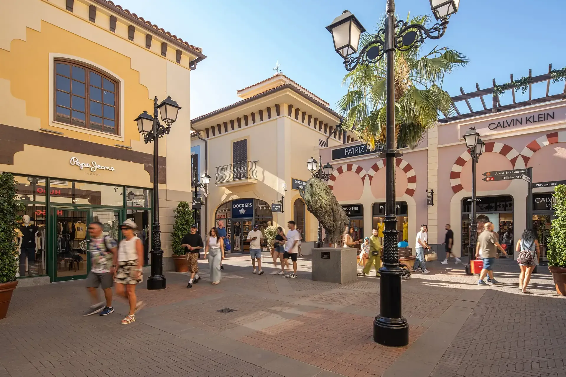 Outdoor shopping plaza with storefronts, pedestrians, and palm trees under a sunny sky