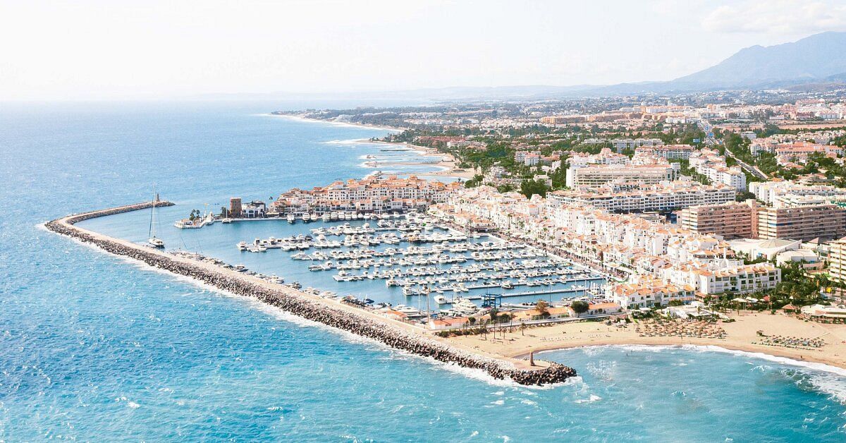 Aerial view of a coastal marina with yachts, a sandy beach, and a seaside town beside blue water.