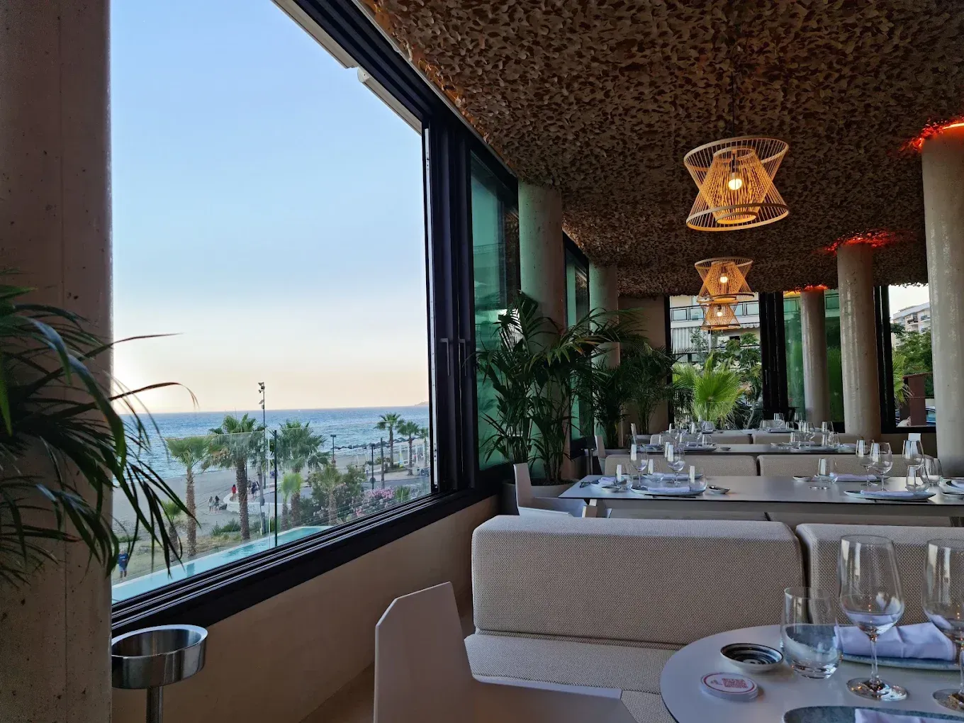 Seaside restaurant dining area with table settings, hanging lights, and ocean view through large windows