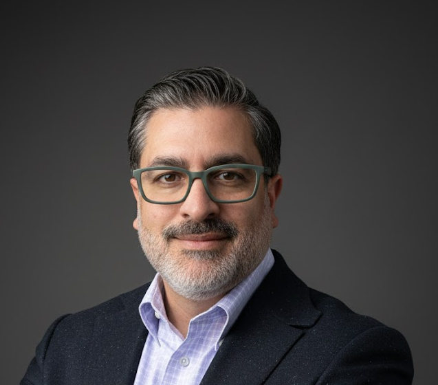 Studio headshot of a smiling person in glasses and a blue blazer 