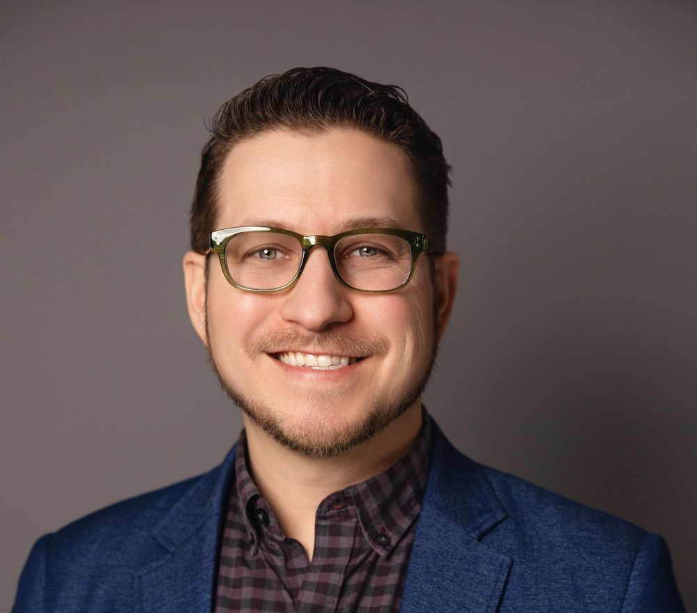 Studio headshot of a smiling person in glasses and a blue blazer on a gray background