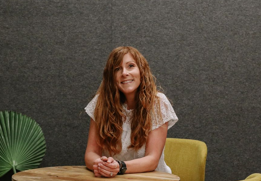 Woman seated at a table, smiling against a gray wall with a green chair and fan-shaped decor.