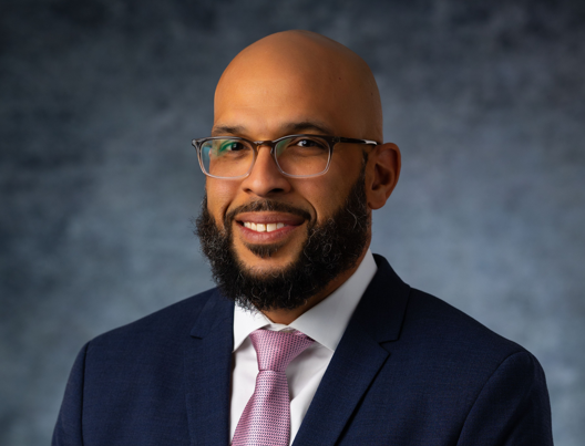 Studio portrait of a smiling man in glasses, navy suit, and pink tie against a gray background