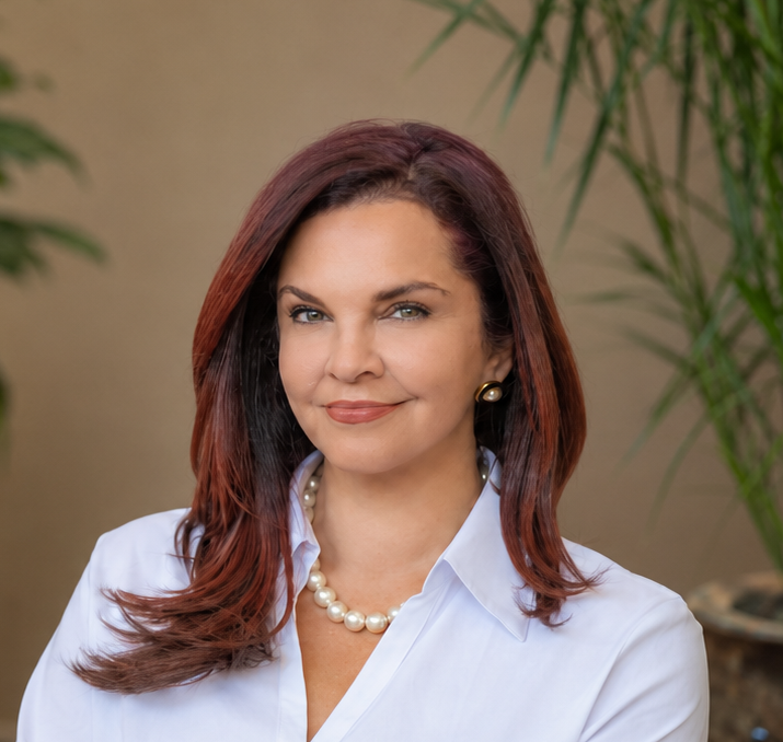 Woman standing against a white background, wearing a white blazer and gold necklace, facing forward and smiling.