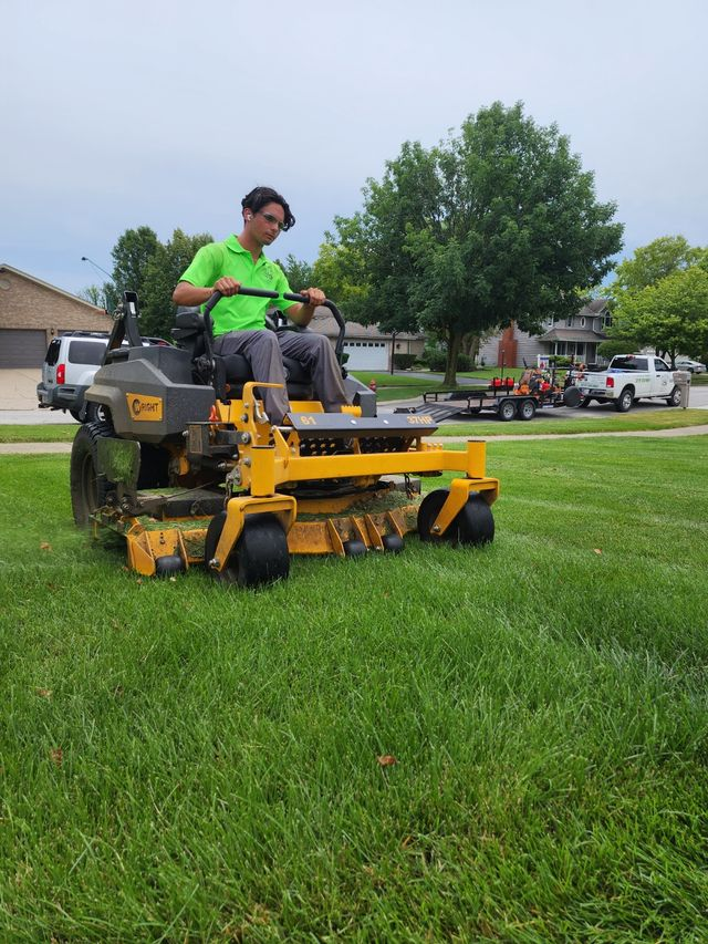Man riding a yellow lawn mower on a grassy field near parked cars and trees