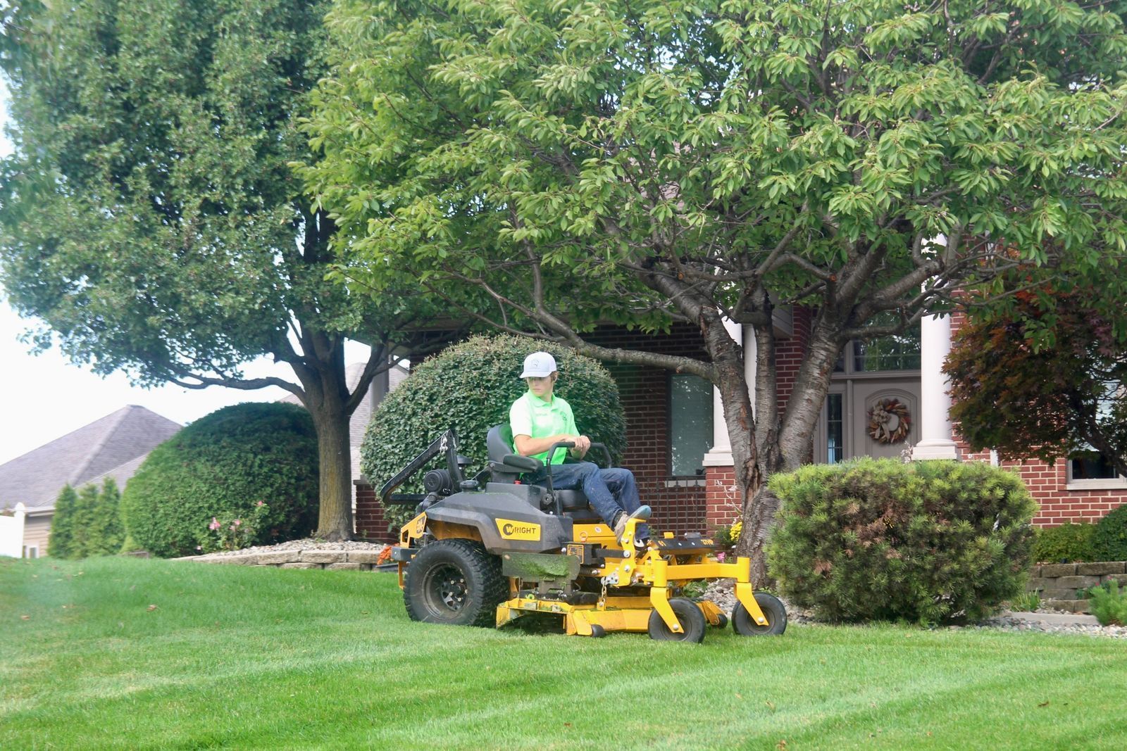 Person riding a yellow lawn mower on a green lawn beside a house and trees