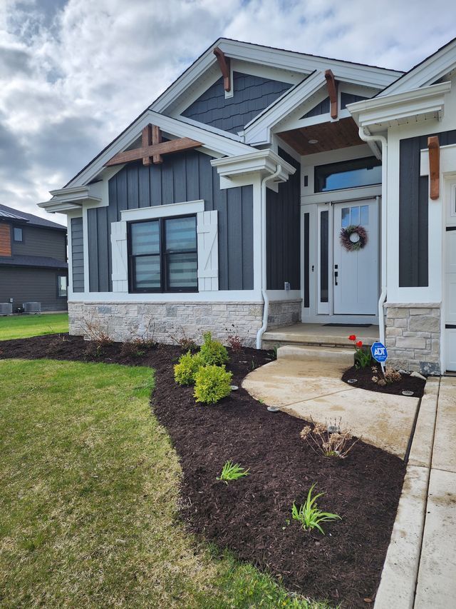 Front yard of a house with dark siding, white trim, stone base, and a mulched flower bed along the walkway.