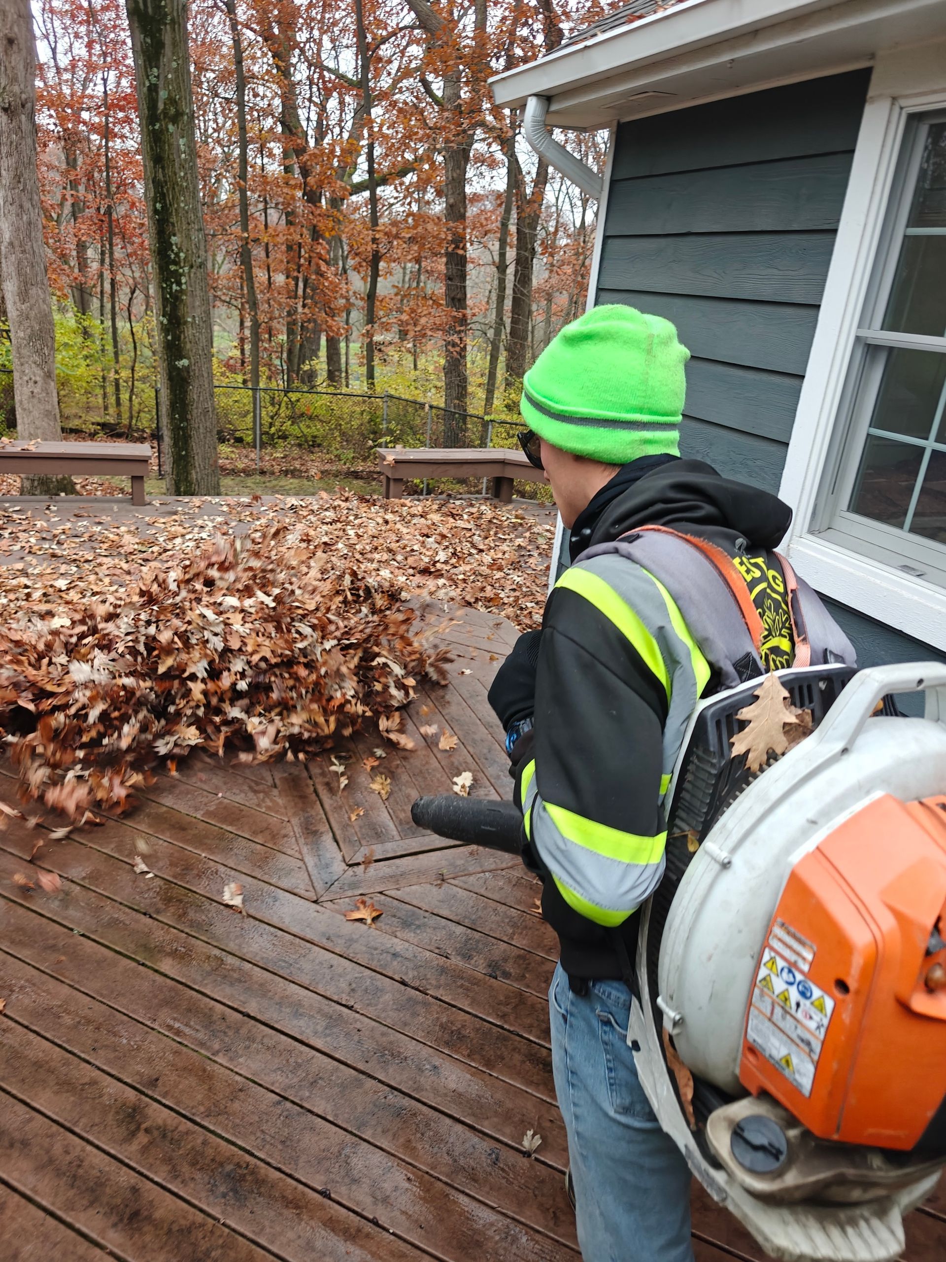 Worker in neon vest and green beanie blowing leaves off a wooden deck beside a house