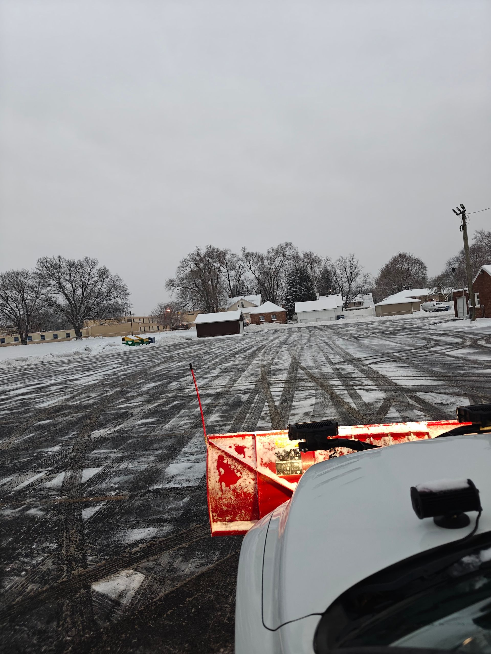 Snowplow clearing a snowy gravel lot, with snow-covered trees and houses in the background.