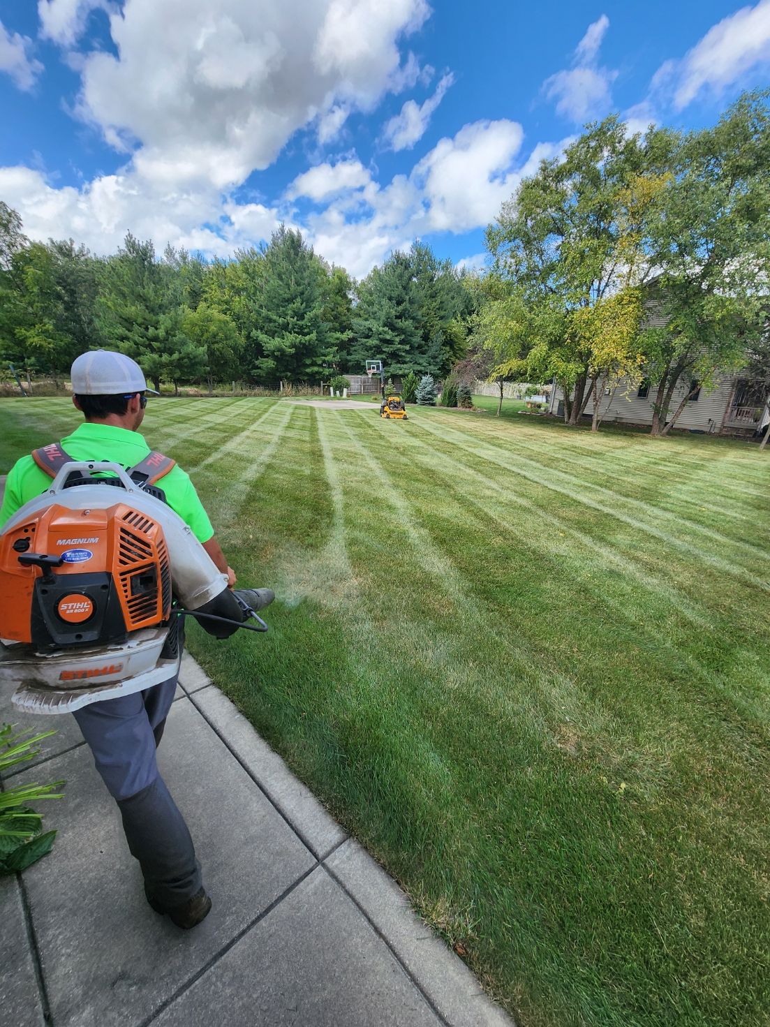 Worker using a backpack blower trims a green lawn beside a sidewalk under a blue sky.