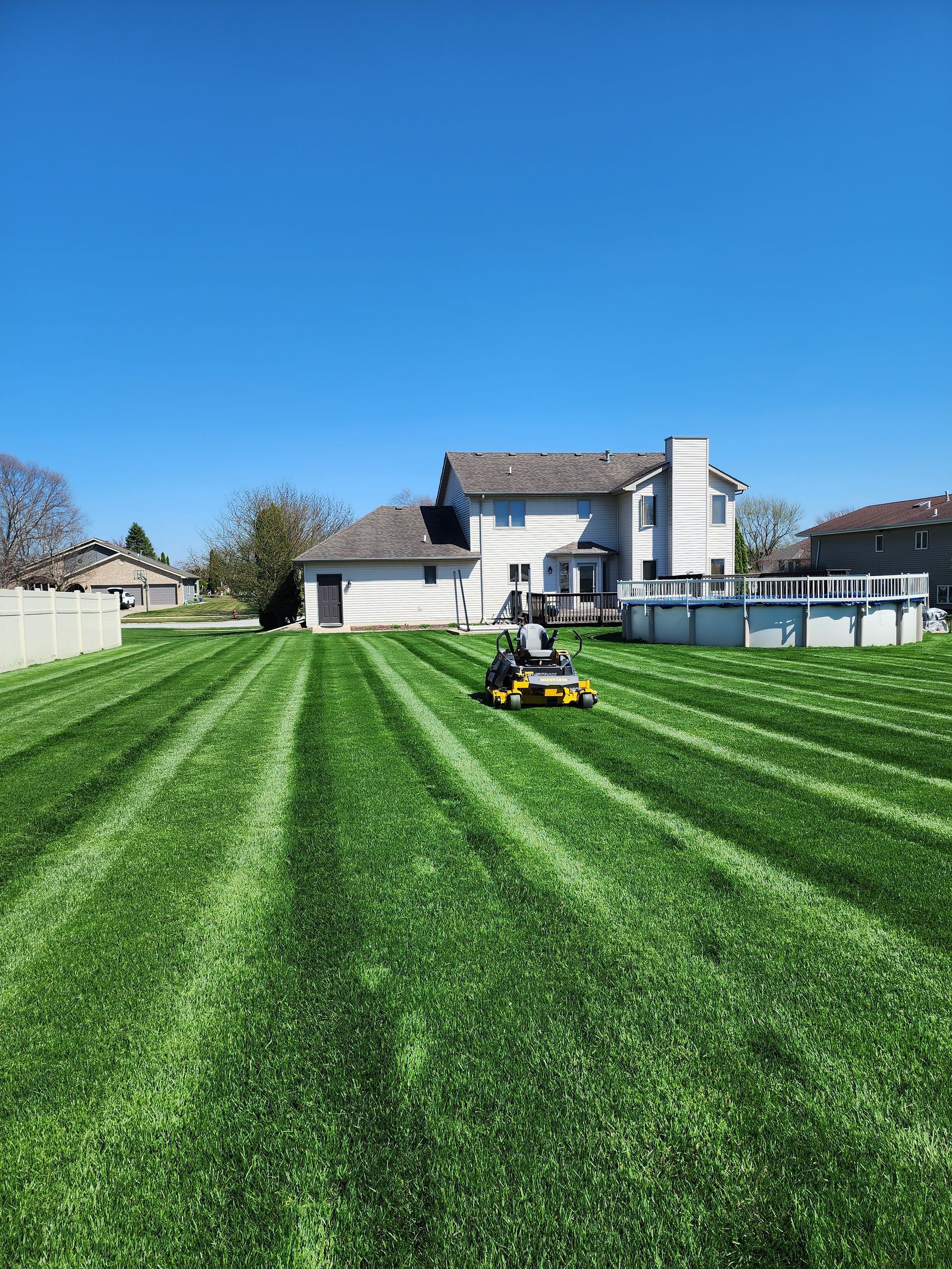 Person mowing a striped green lawn in front of a house under a clear blue sky