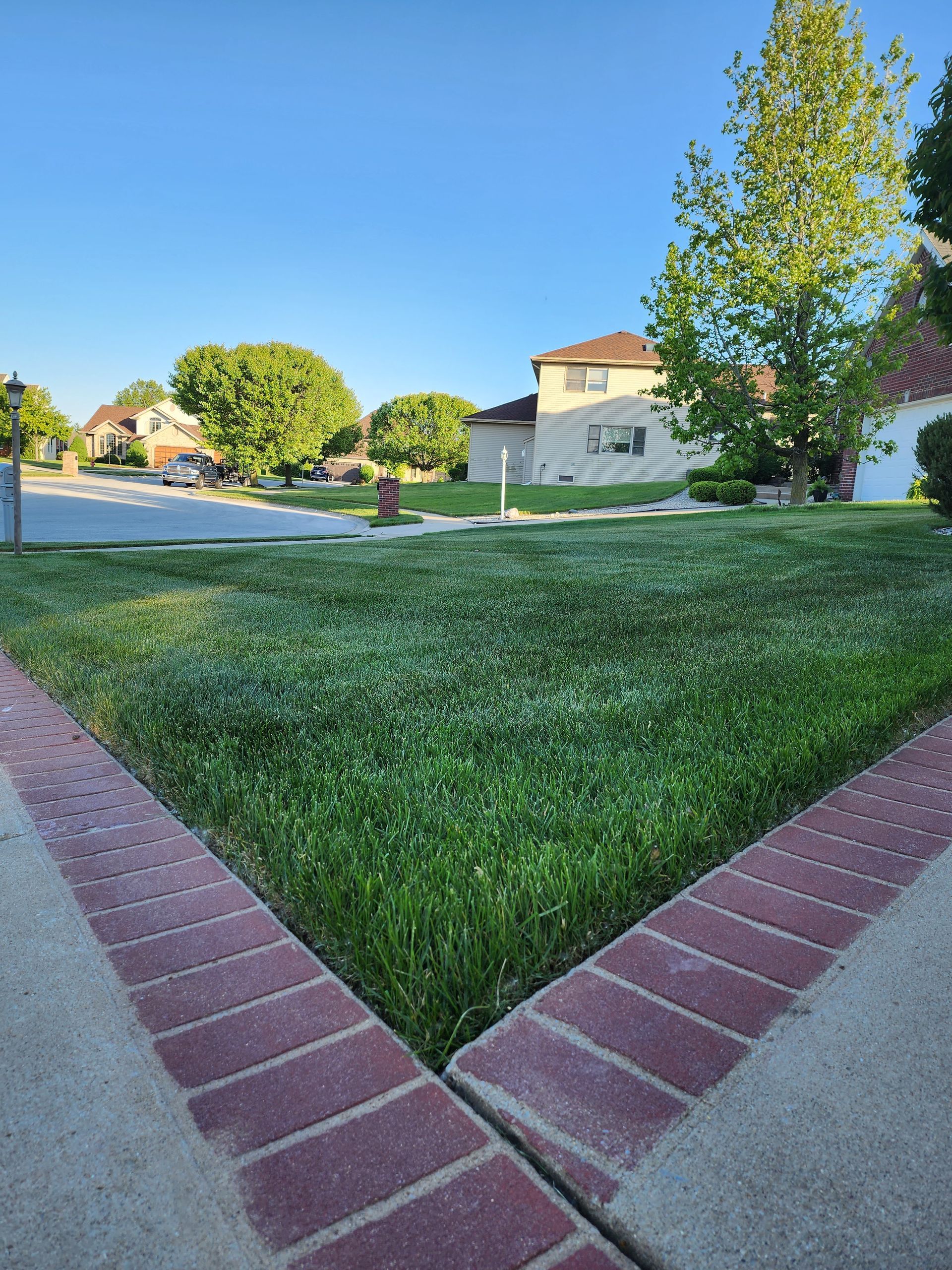 Brick walkways framing a grassy lawn in a sunny suburban park with houses and trees nearby
