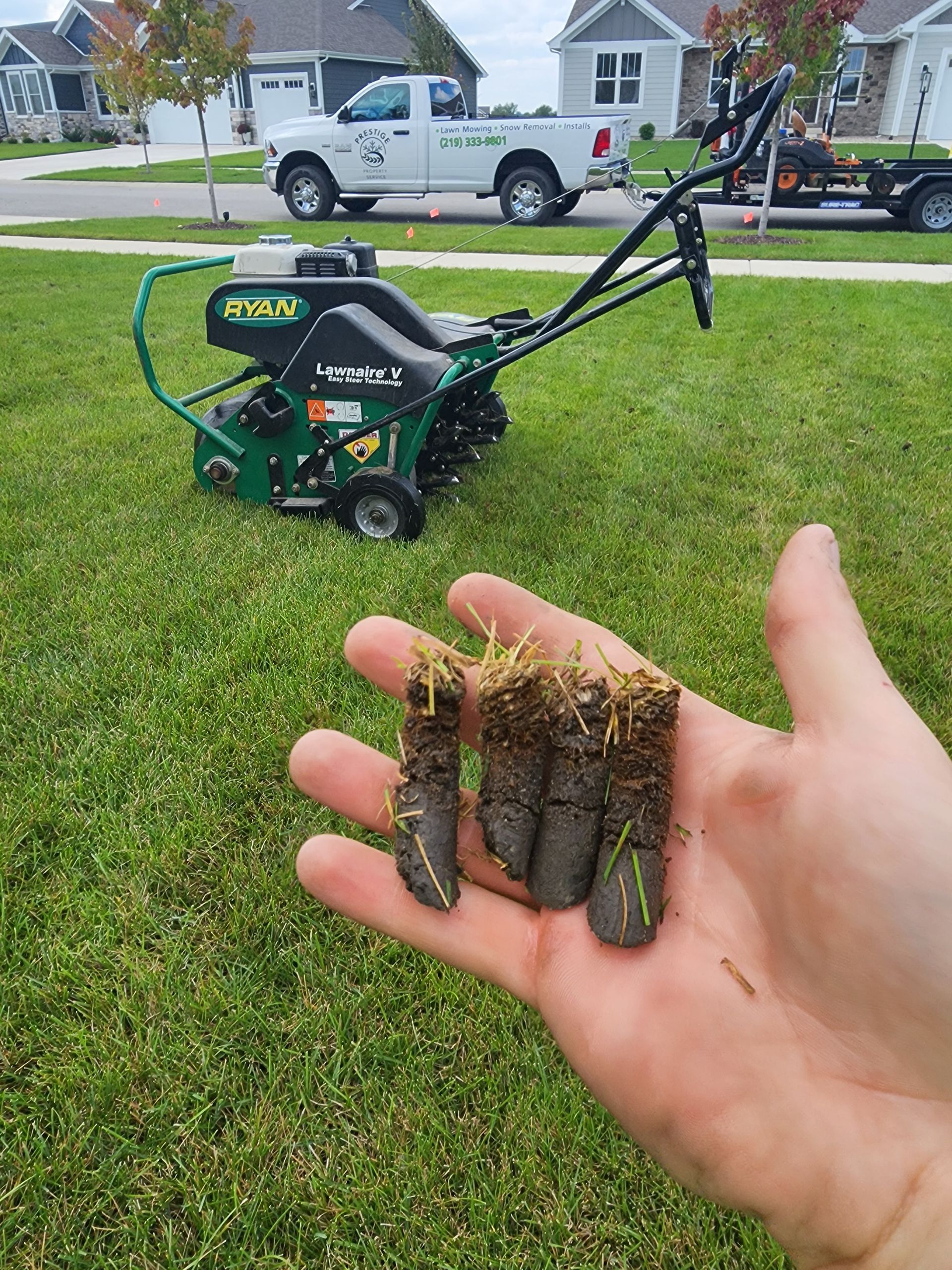 Hand holding soil and grass clumps in front of a green lawn aerator on a suburban lawn