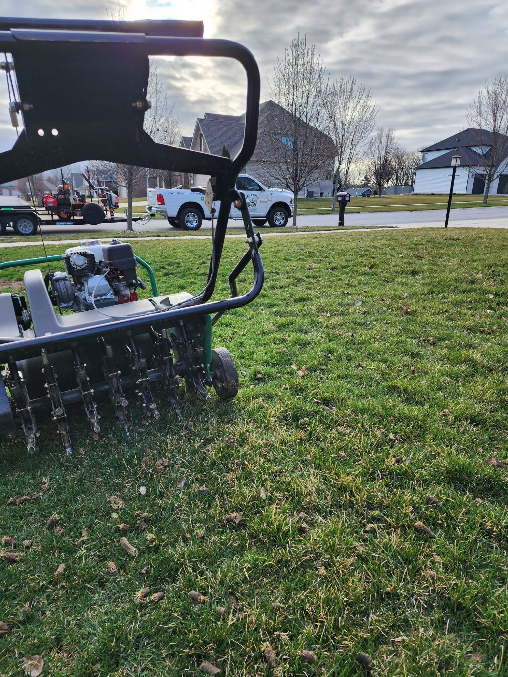 Person mowing a grassy park with a riding mower near parked cars on a sunny day