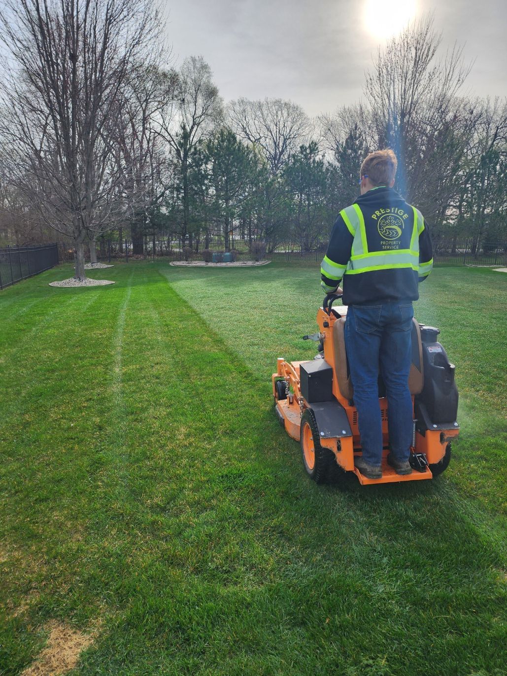 Worker mowing a grassy park with an orange ride-on mower, wearing a hi-vis vest.