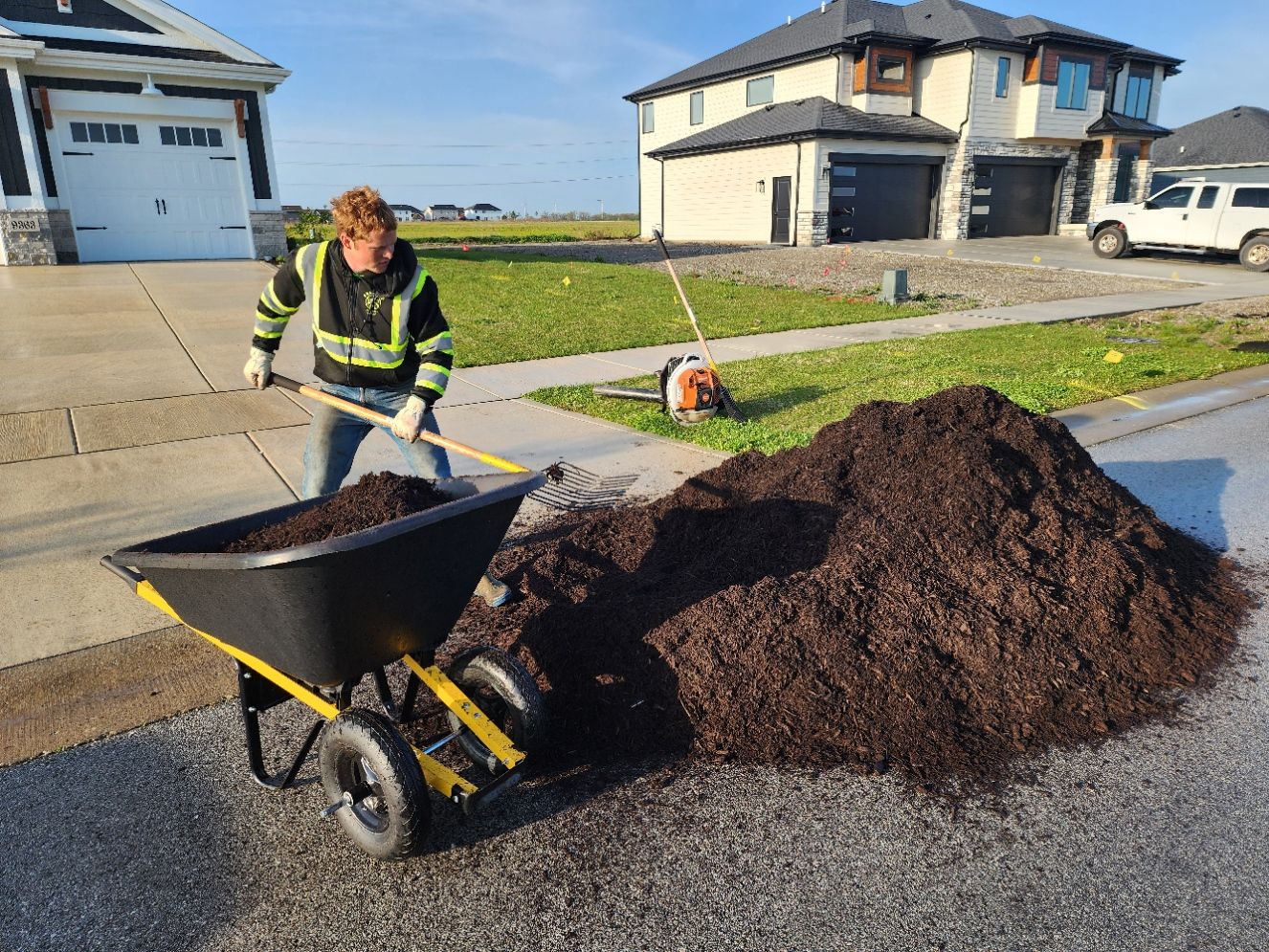 Worker shovels asphalt into a wheelbarrow beside a large pile on a residential street.