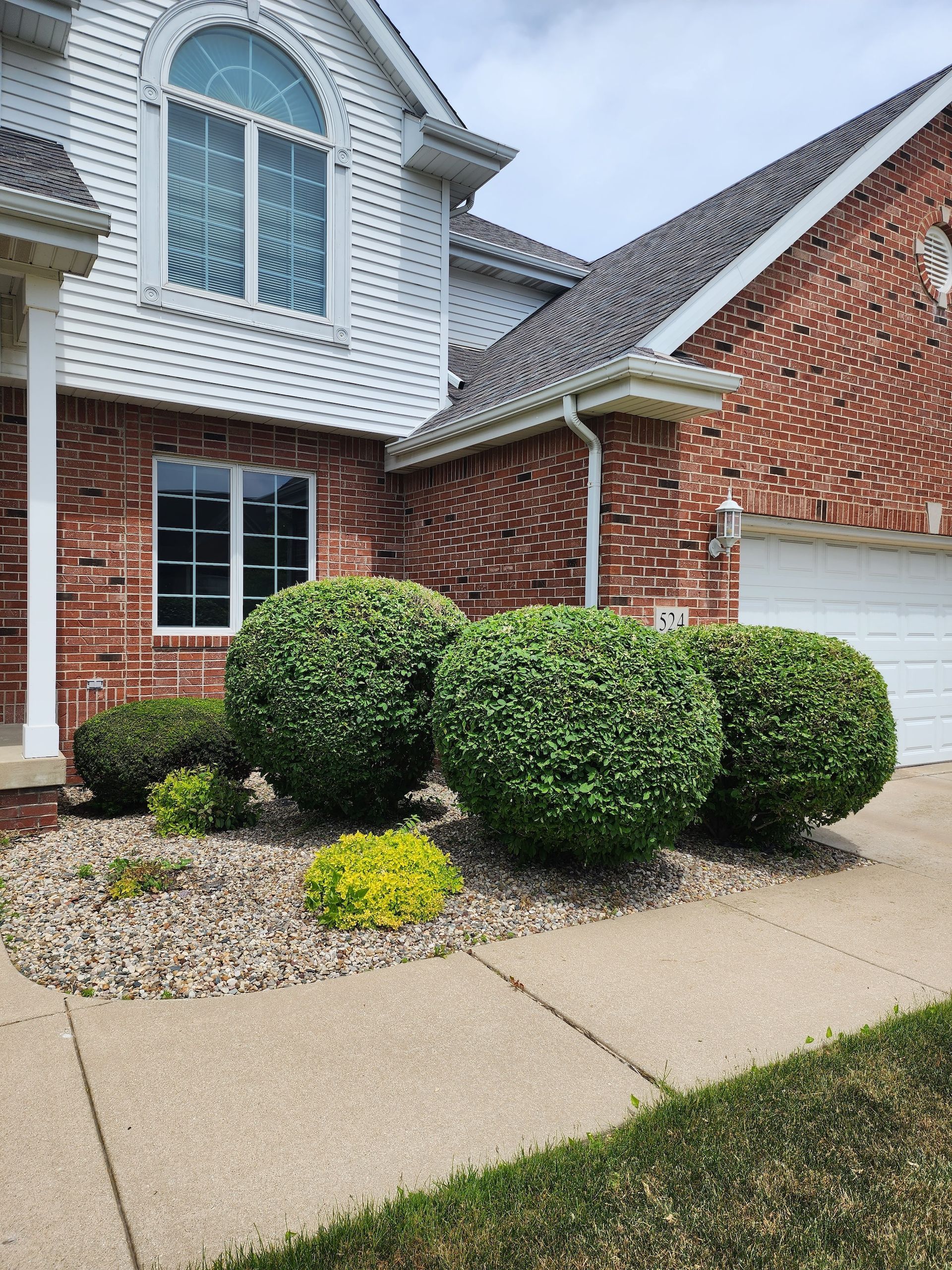 Front of a brick house with white siding, trimmed bushes, and a concrete walkway.