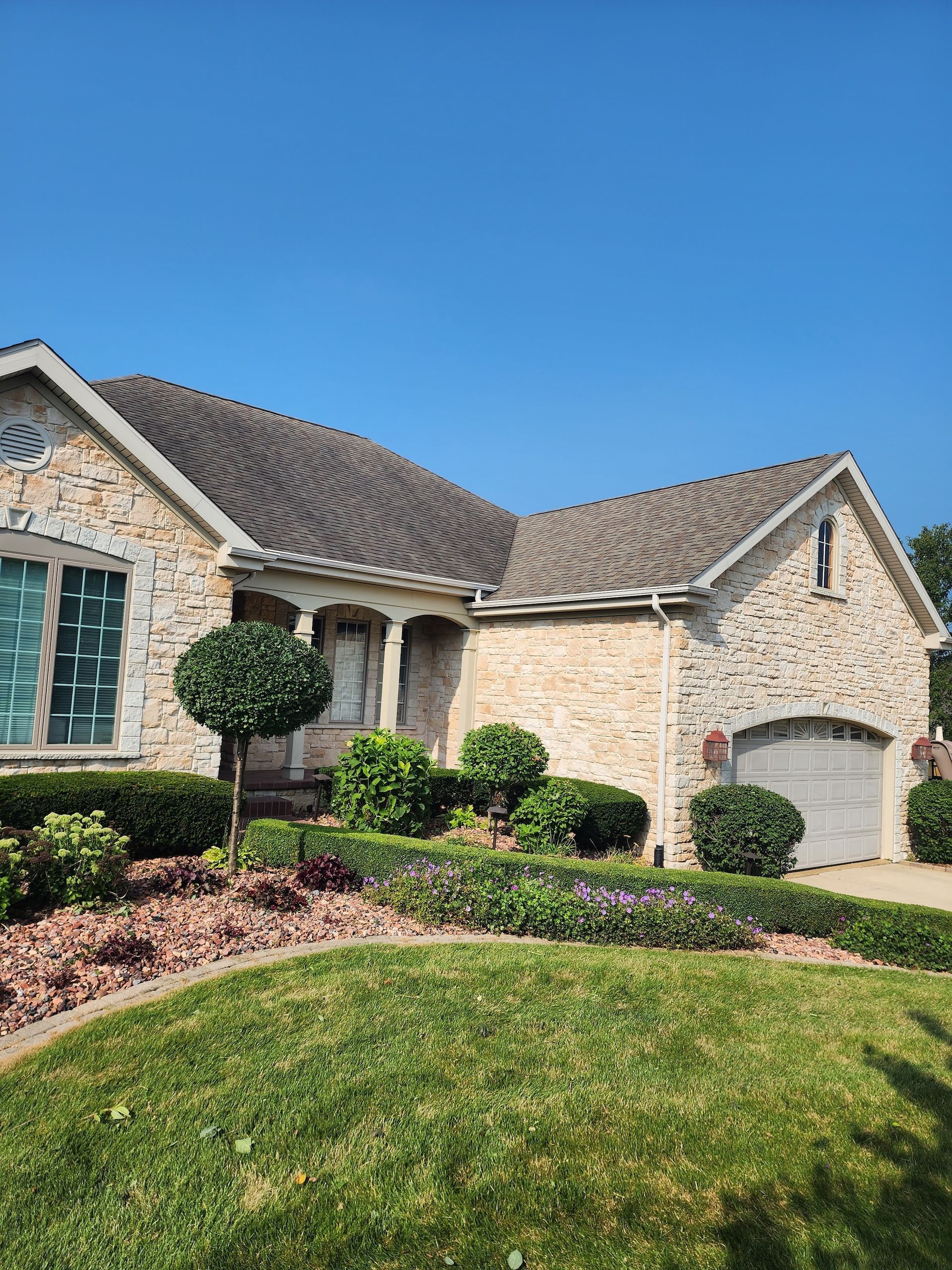 Stone suburban house with gray roof, green lawn, manicured shrubs, and garage under a blue sky