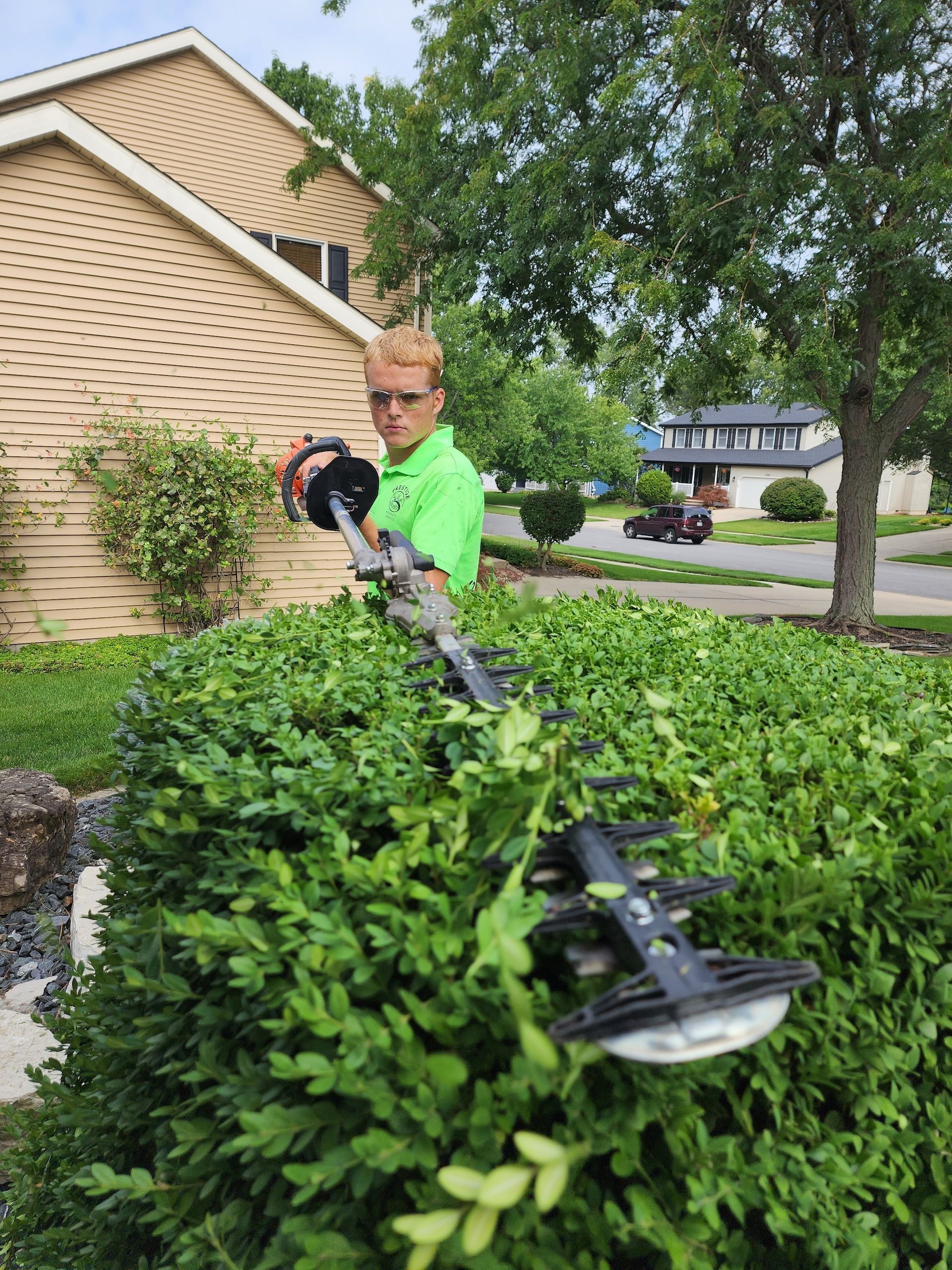 Person in neon green shirt trimming hedges in a front yard near a house.