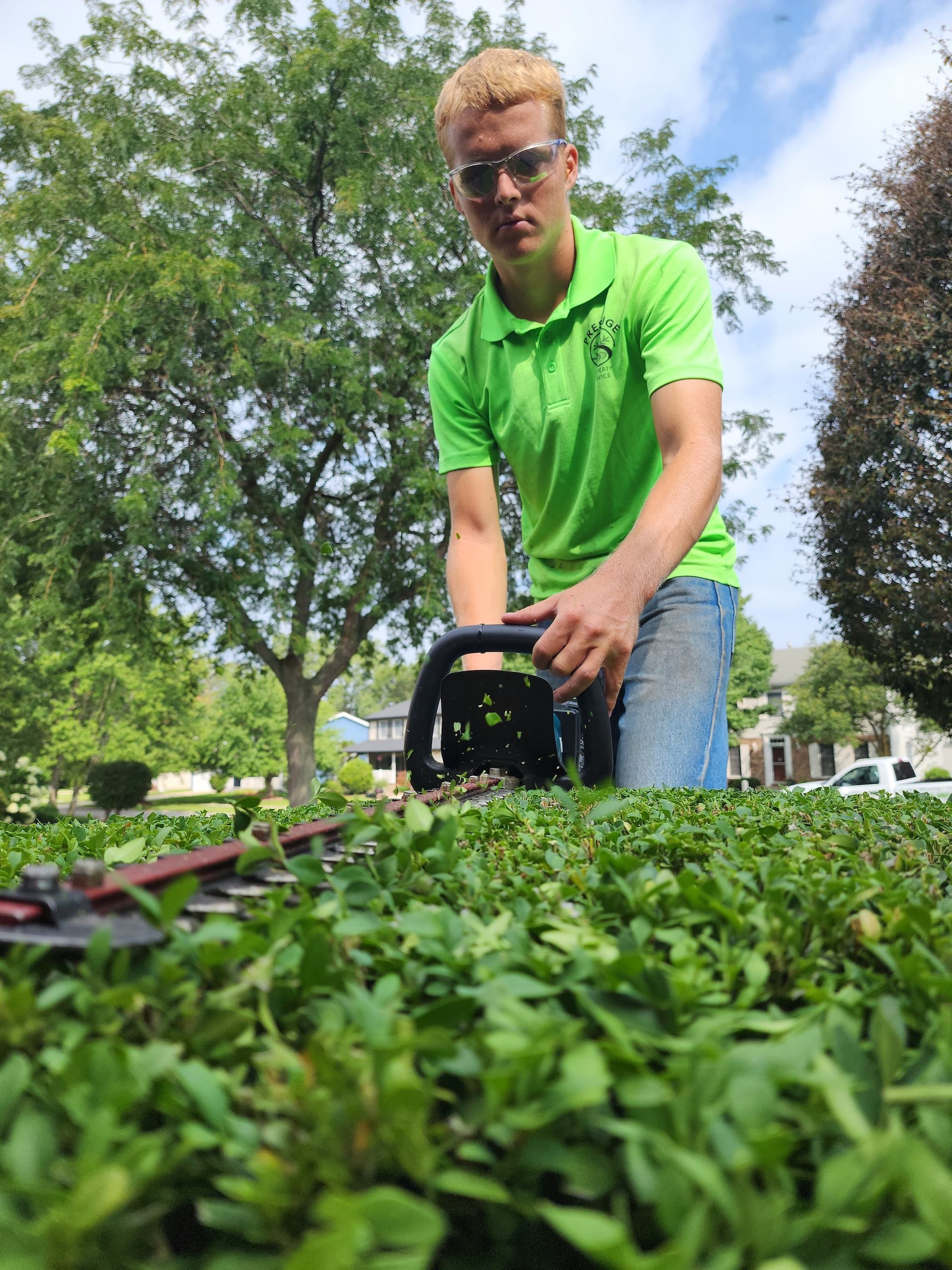 Person in a bright green shirt pushing a lawn mower in a grassy yard with trees in the background