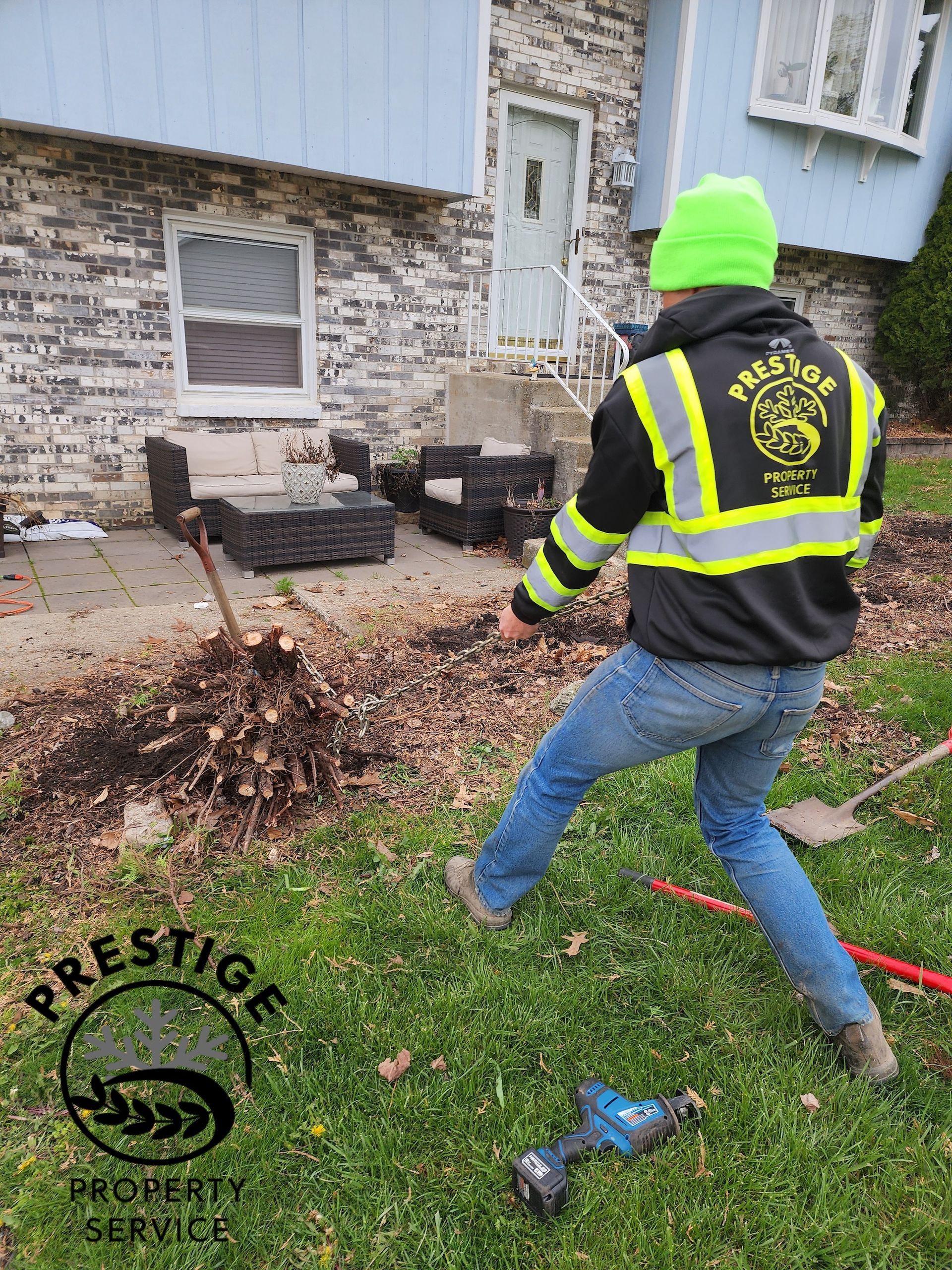 Worker in neon beanie and safety vest using a blower to clear leaves beside a house entrance.