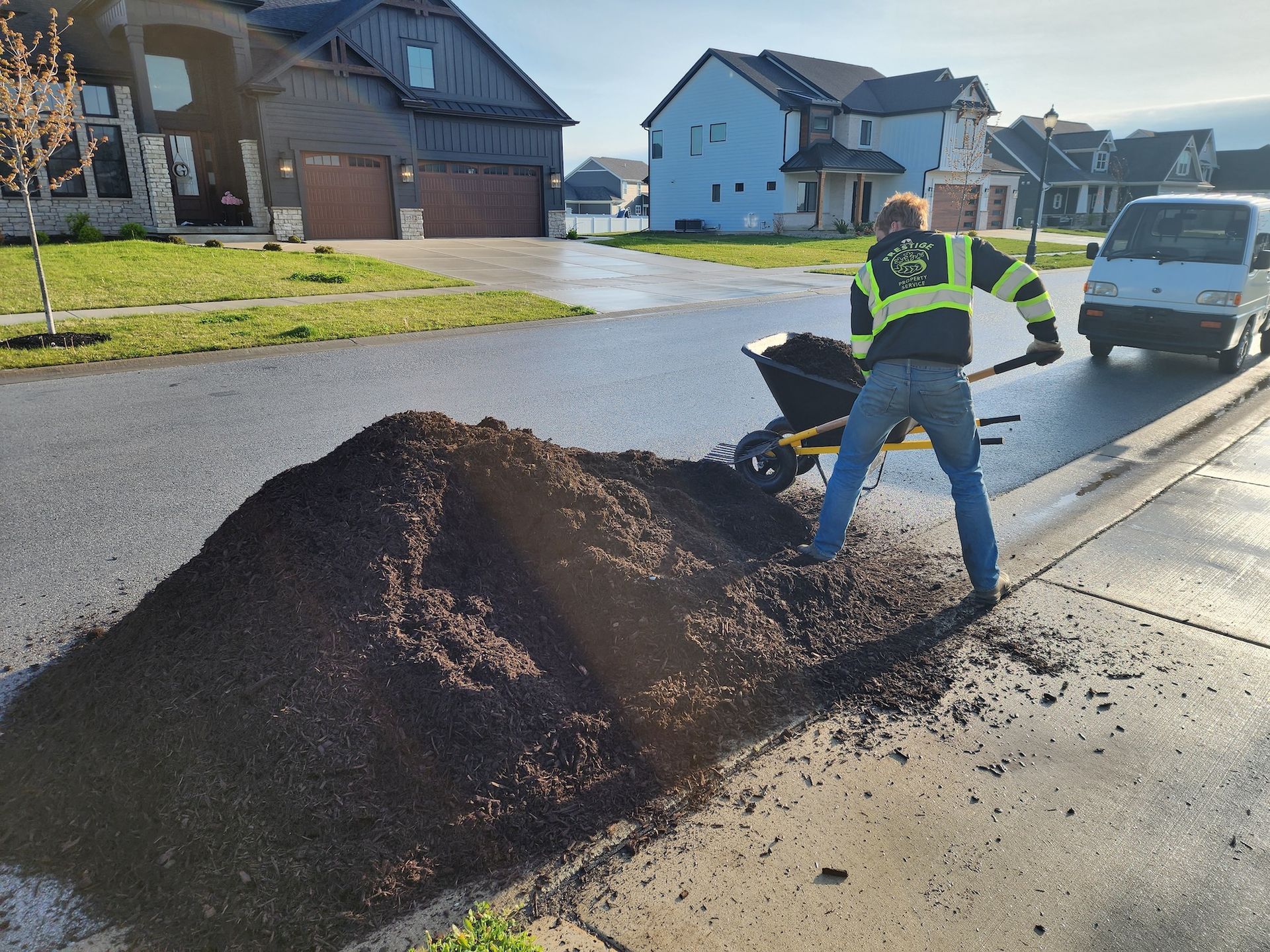 Worker spreading a large pile of dark mulch on a suburban street beside parked cars and houses