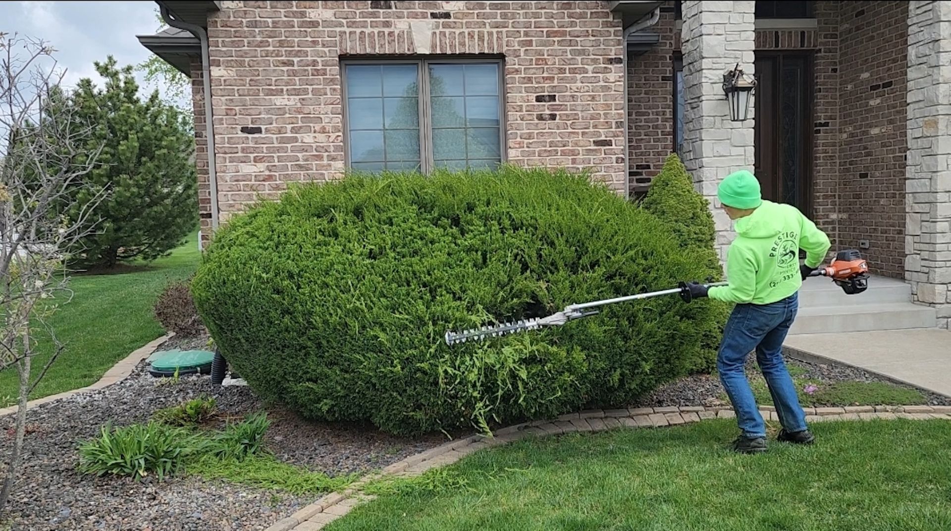 Person in neon green jacket trimming a large shrub beside a brick house with a hedge trimmer