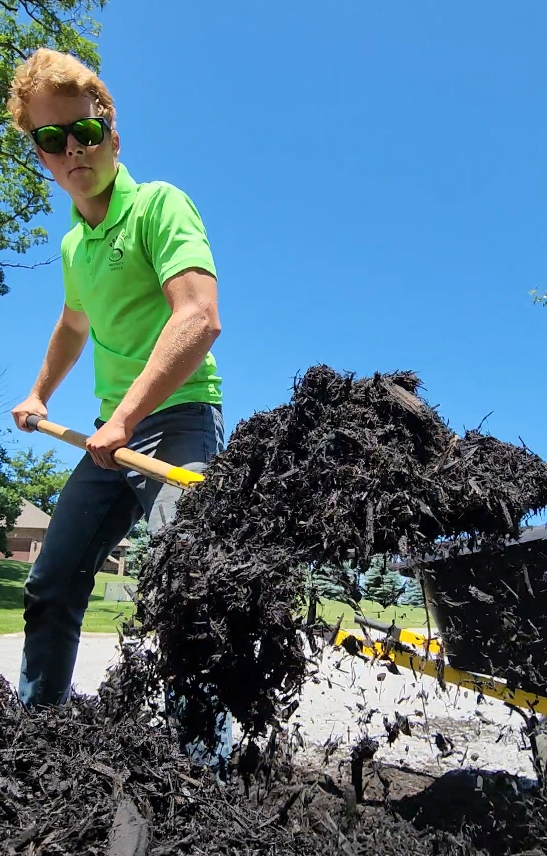 Person in a green shirt shovels dark mulch into a wheelbarrow outdoors under a blue sky