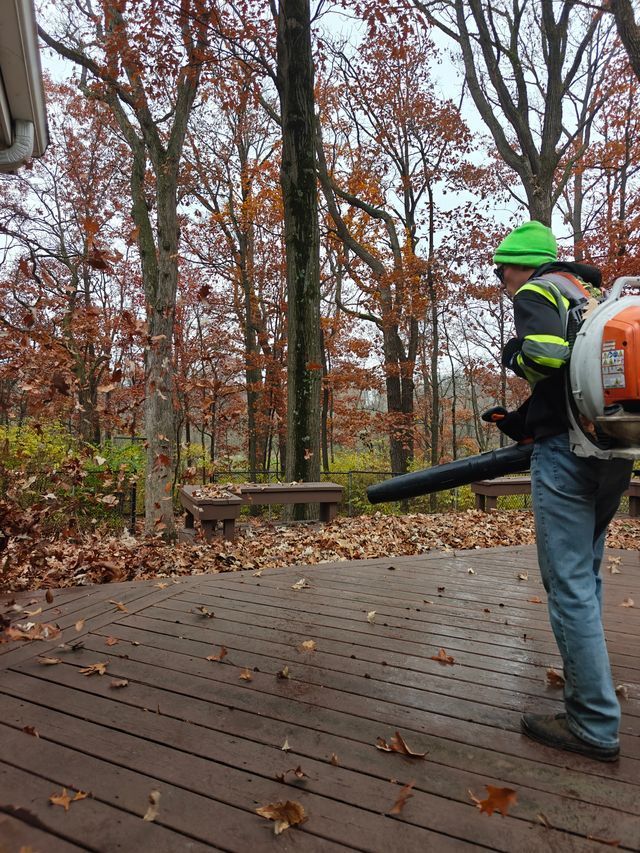 Worker using a leaf blower on a wooden deck in a wooded backyard during fall