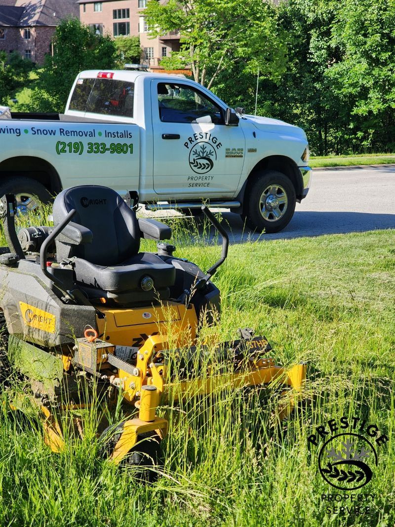 Yellow lawn mower in tall grass with a white utility truck parked behind it on a residential street.