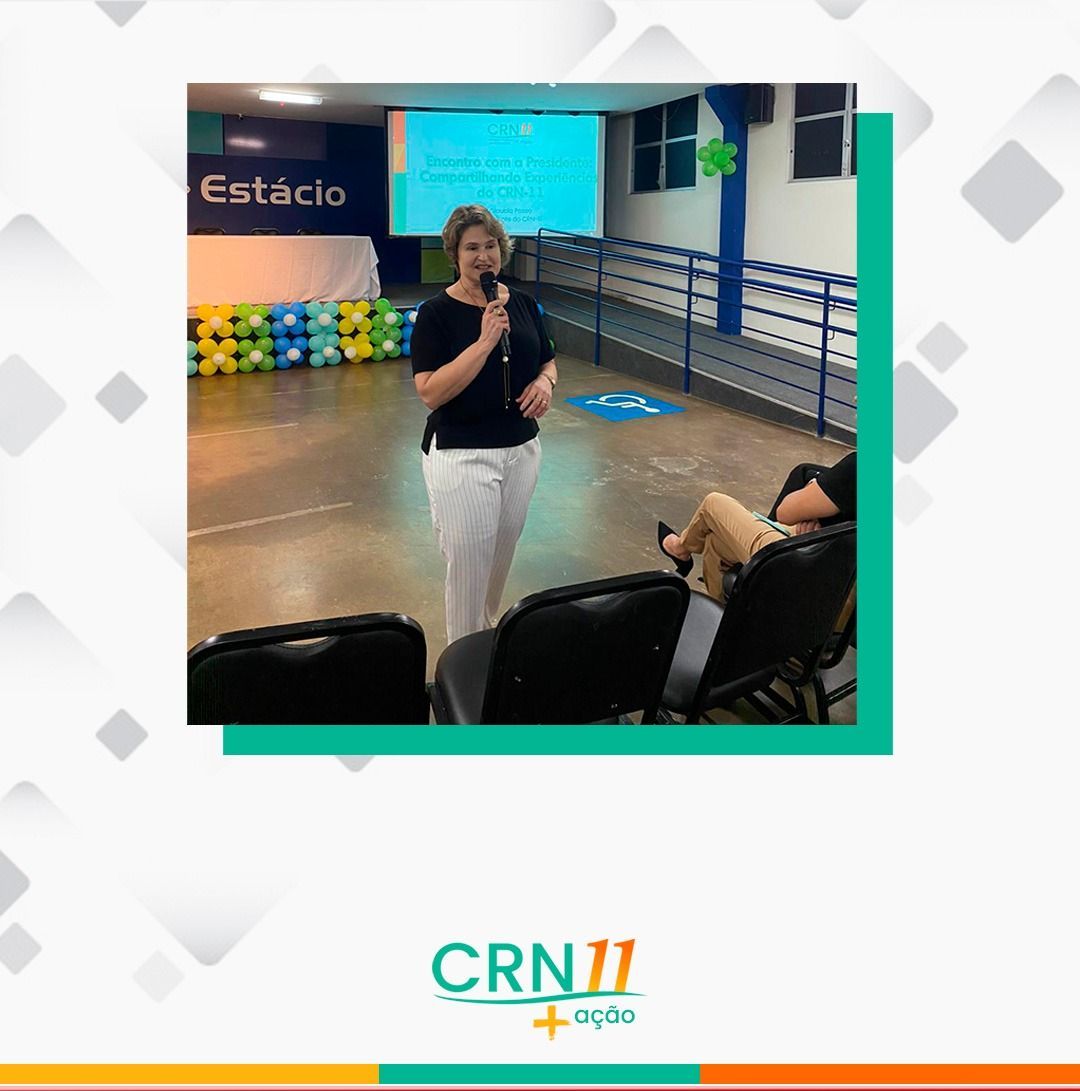 Mulher falando ao microfone em um auditório, em pé perto de um palco azul com espectadores sentados.