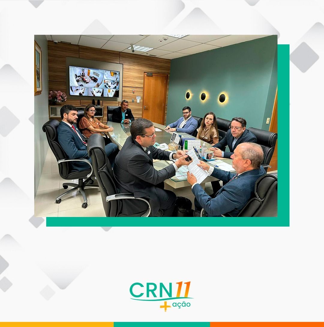 Pessoas em uma sala de reuniões discutindo documentos em torno de uma mesa com um painel na parede.