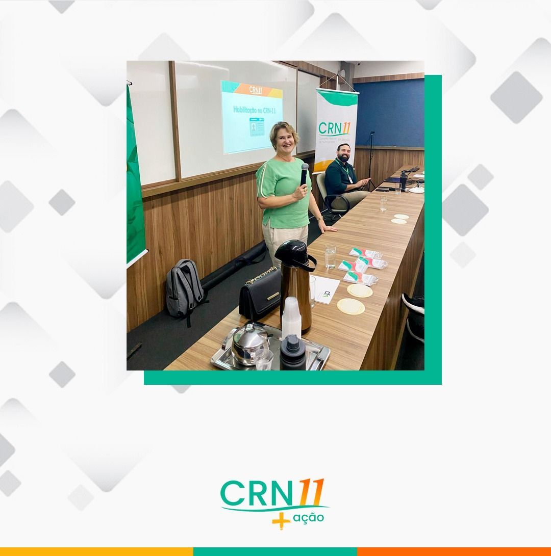 Palestrante apresentando em uma mesa de conferência com slides projetados atrás dela em uma sala de reuniões.