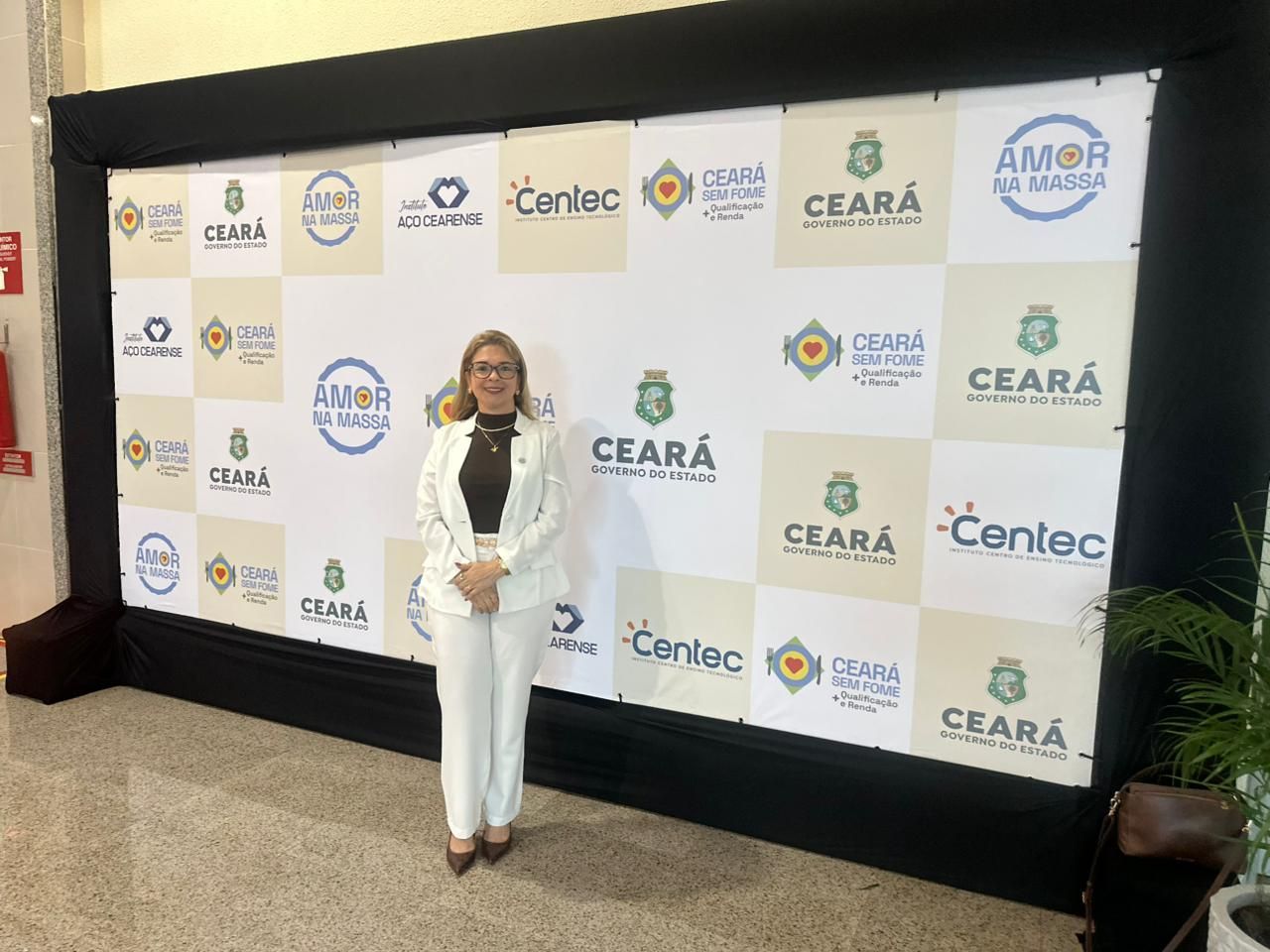 Mulher de terno branco em pé diante de um painel com o logotipo de um patrocinador em um evento interno.