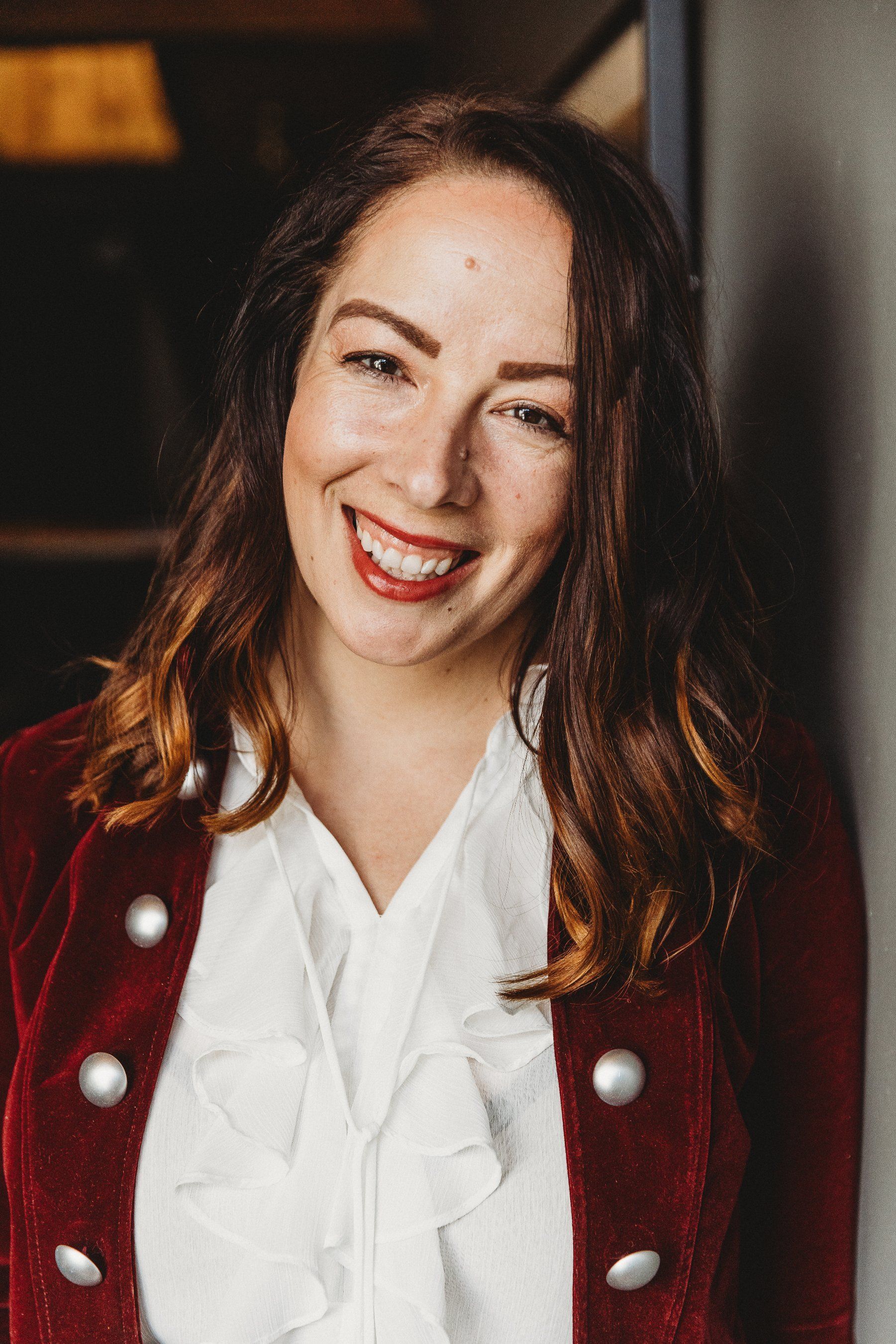 Woman smiling widely, wearing a red jacket over a white blouse, dark hair, indoors.
