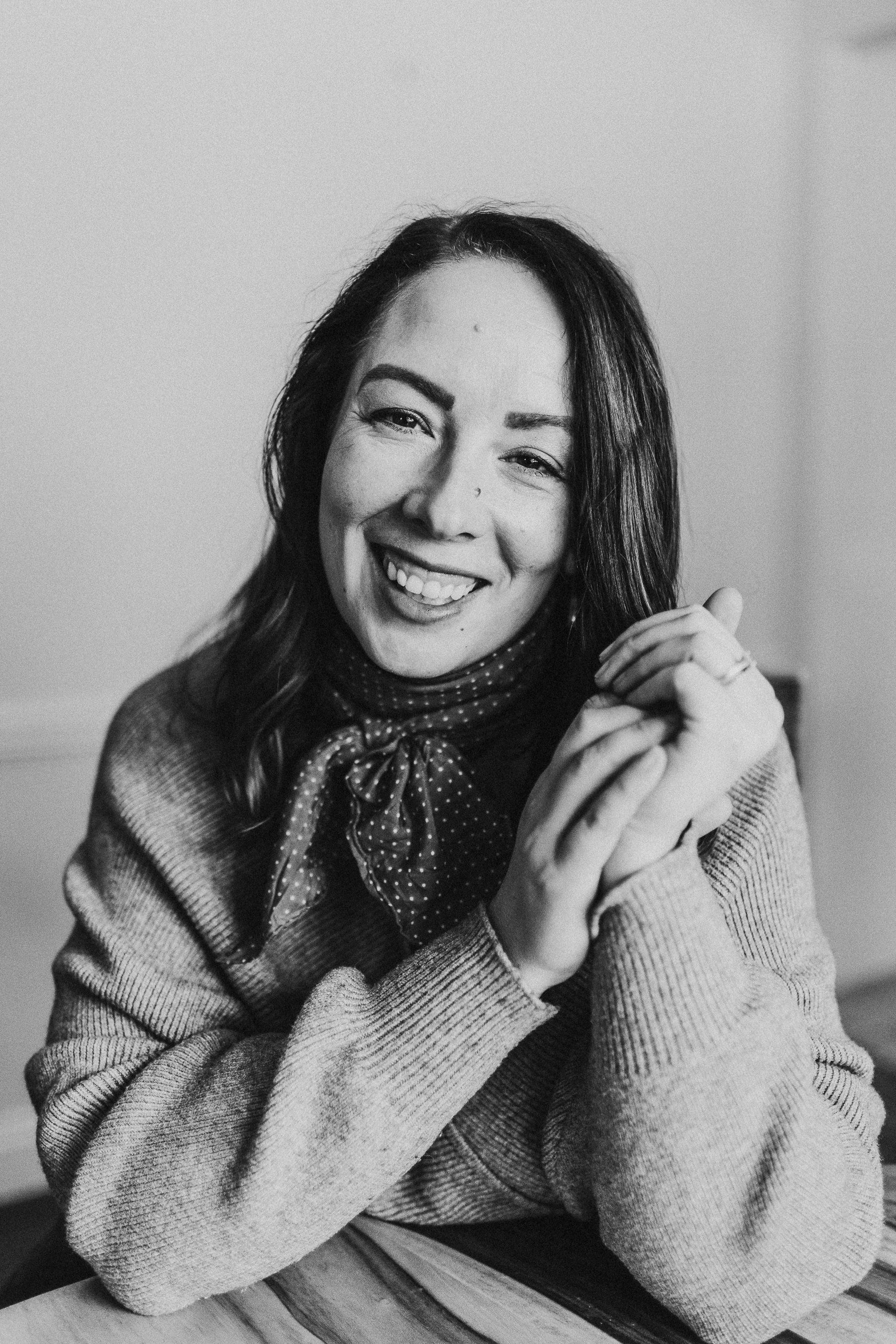 Woman smiles at the camera, arms crossed on a table. She wears a sweater and scarf.