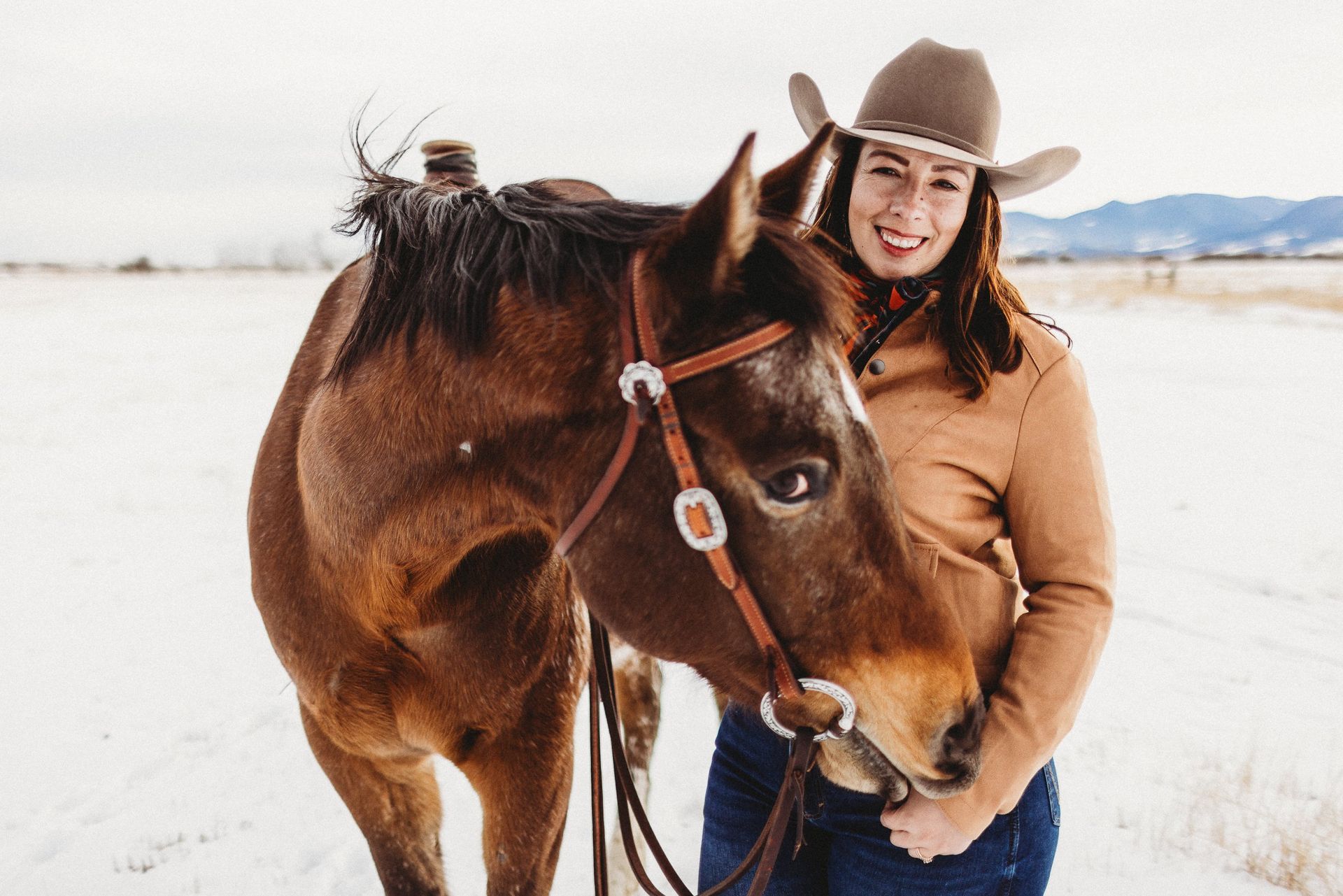 Woman in cowboy hat smiles with brown horse in snowy field.