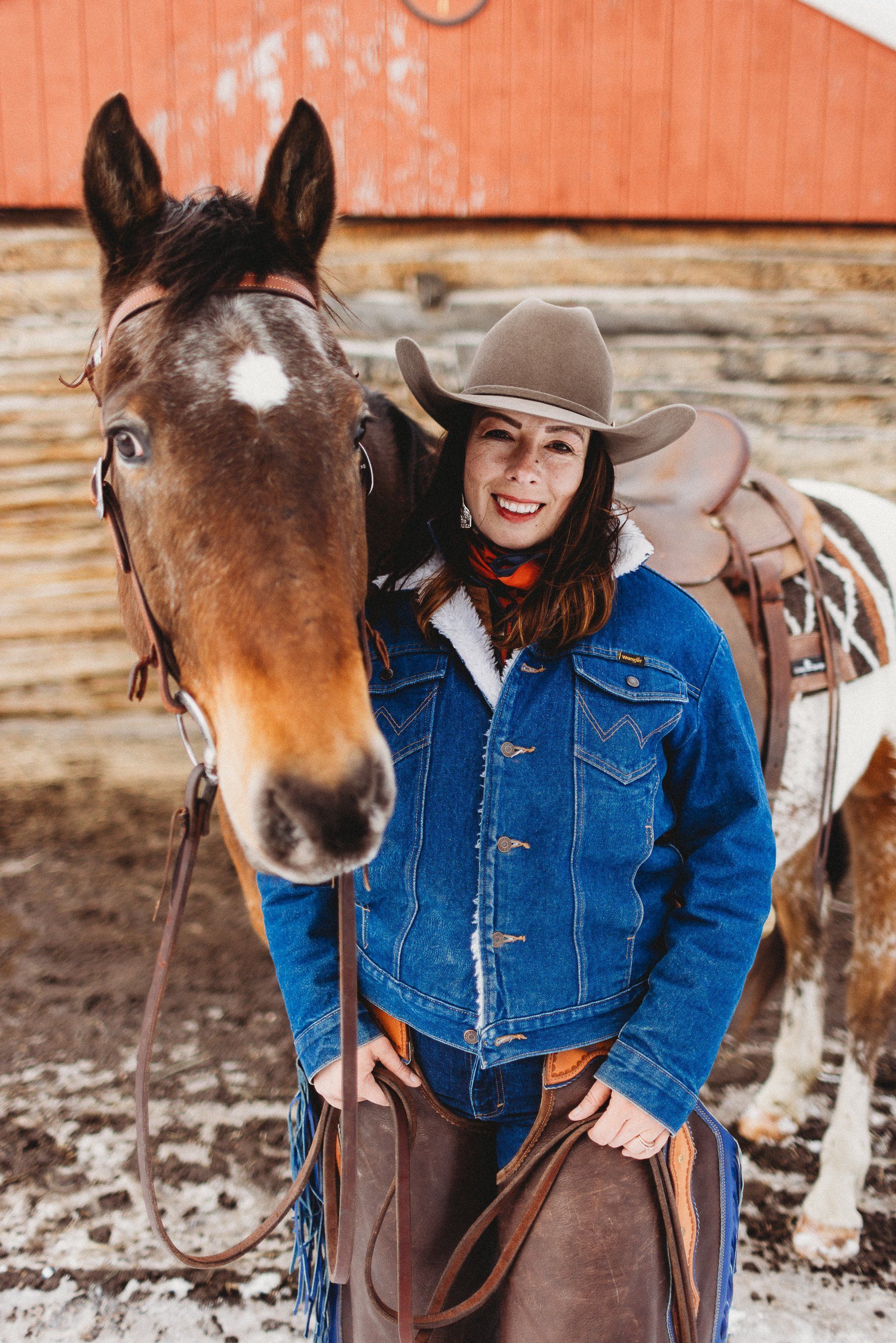 Woman in cowboy hat and denim jacket poses with a horse in front of a red barn.